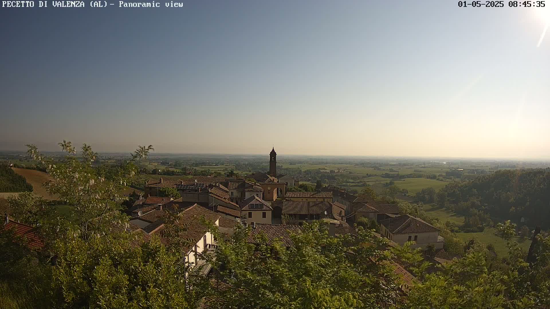 A panoramic view shows a small village nestled among green hills and fields under a clear, bright sky.