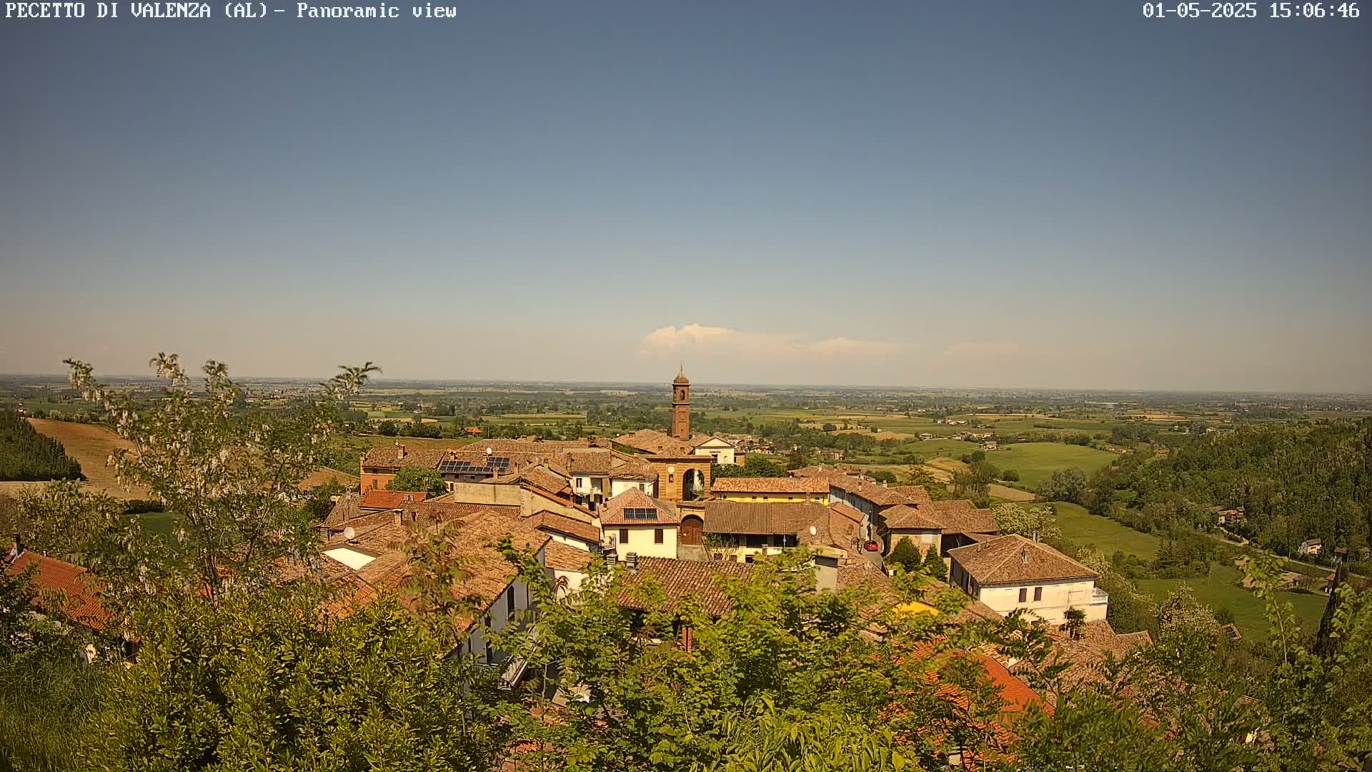 A panoramic view of a village with red-tiled roofs, a tall bell tower, and surrounding green fields under a clear, sunny sky.