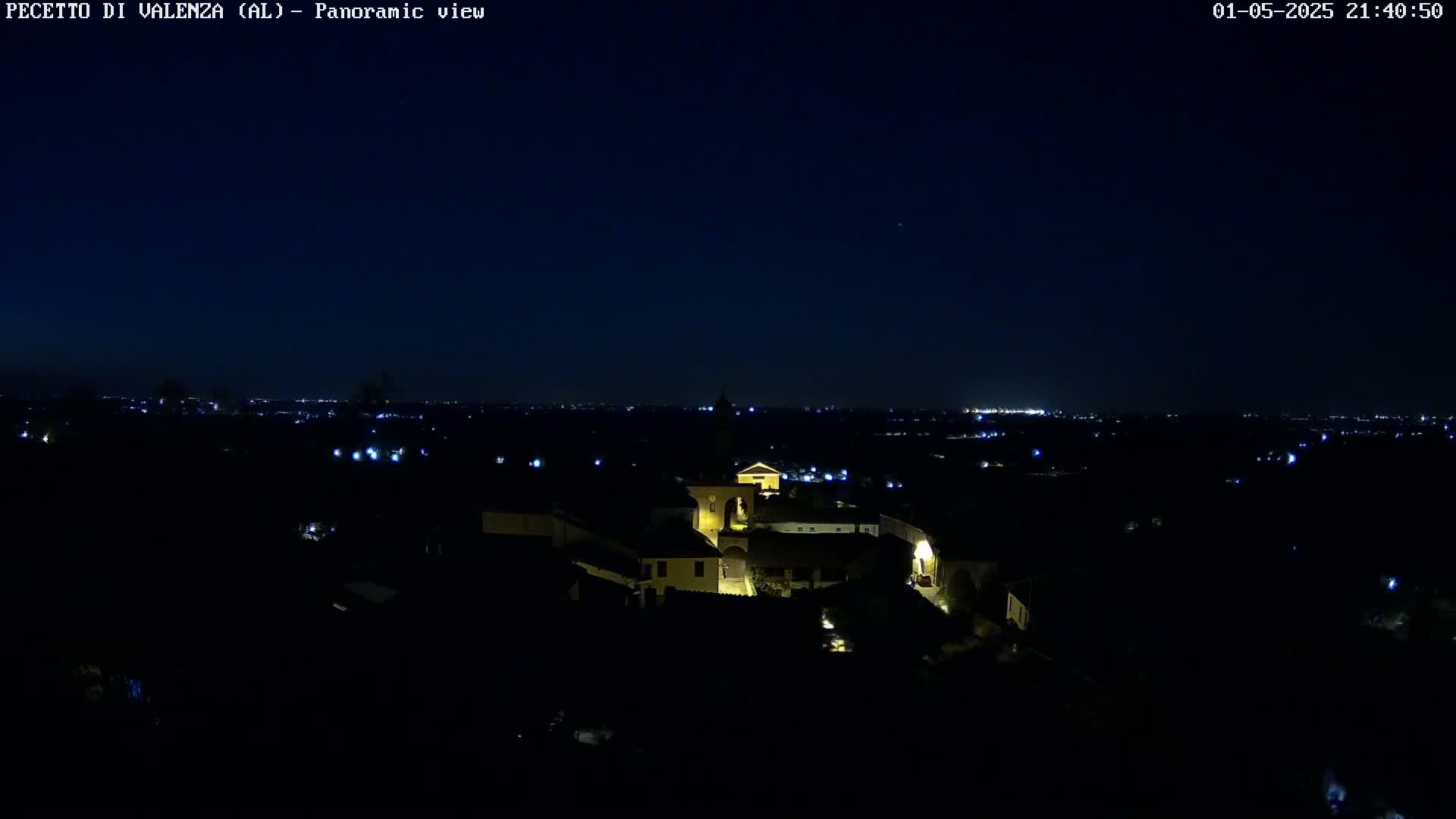 A nighttime panoramic view shows a small, illuminated village nestled in a dark landscape under a clear, dark sky.