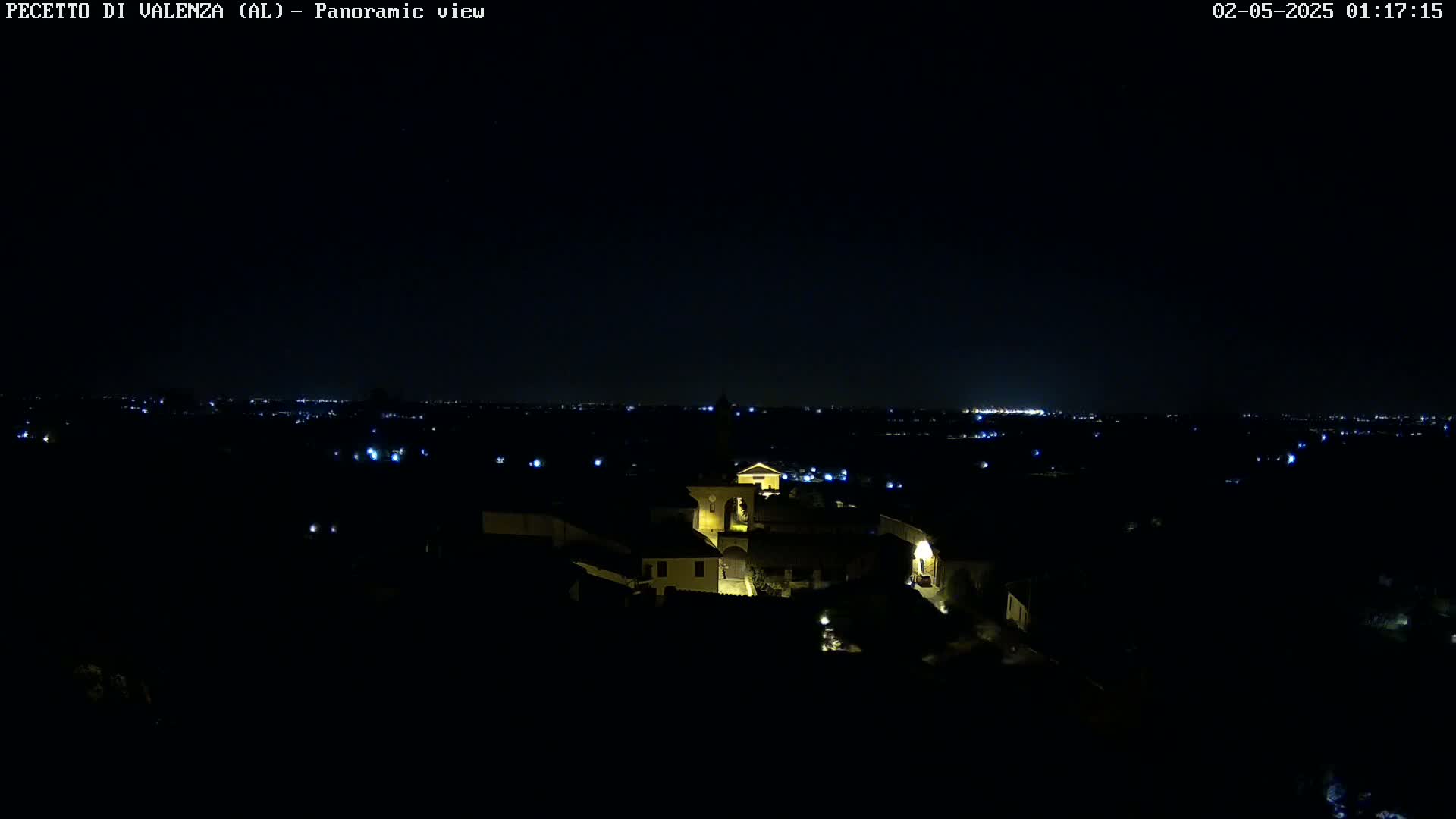 A nighttime, panoramic view shows a small, illuminated village nestled in a dark landscape, with a distant cityscape visible on the horizon.