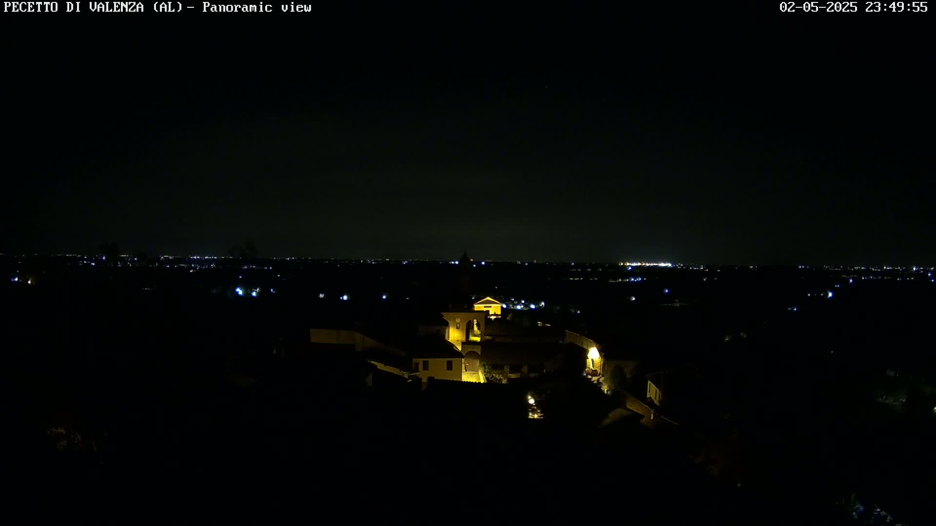 A nighttime panoramic view shows a small, illuminated village nestled in a dark landscape, with a distant city skyline faintly visible in the background under a clear sky.