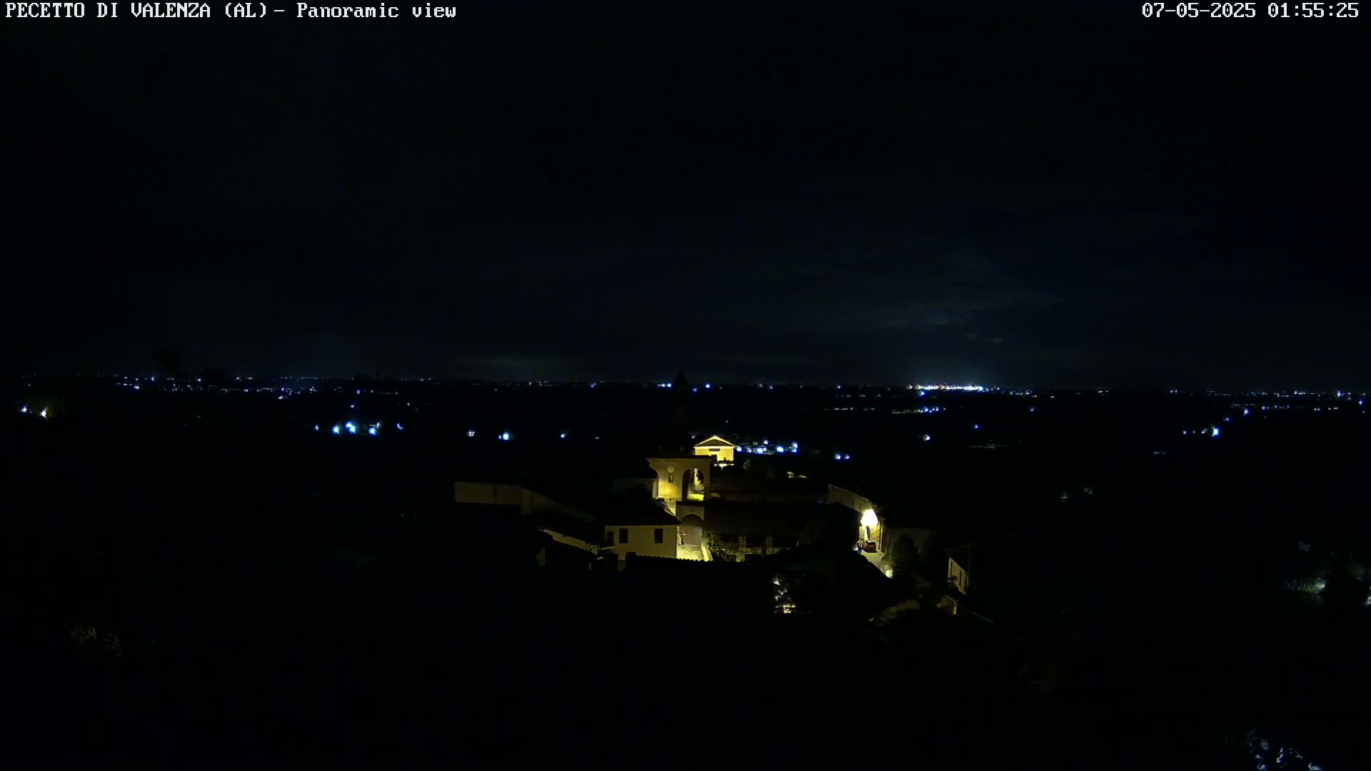 A nighttime panoramic view shows a small, illuminated village nestled in the dark countryside, with a distant city skyline visible on the horizon under a mostly clear sky.