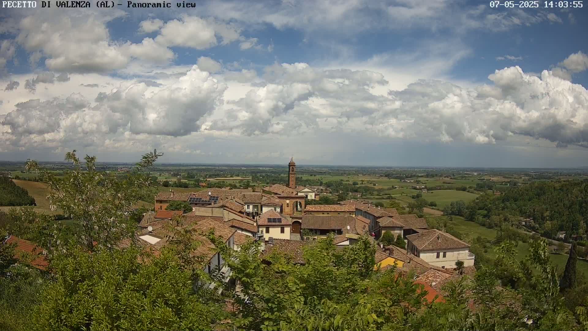 A partly cloudy sky overlooks a small town nestled in a verdant valley, with red-tiled roofs and a tall bell tower visible amongst the buildings.
