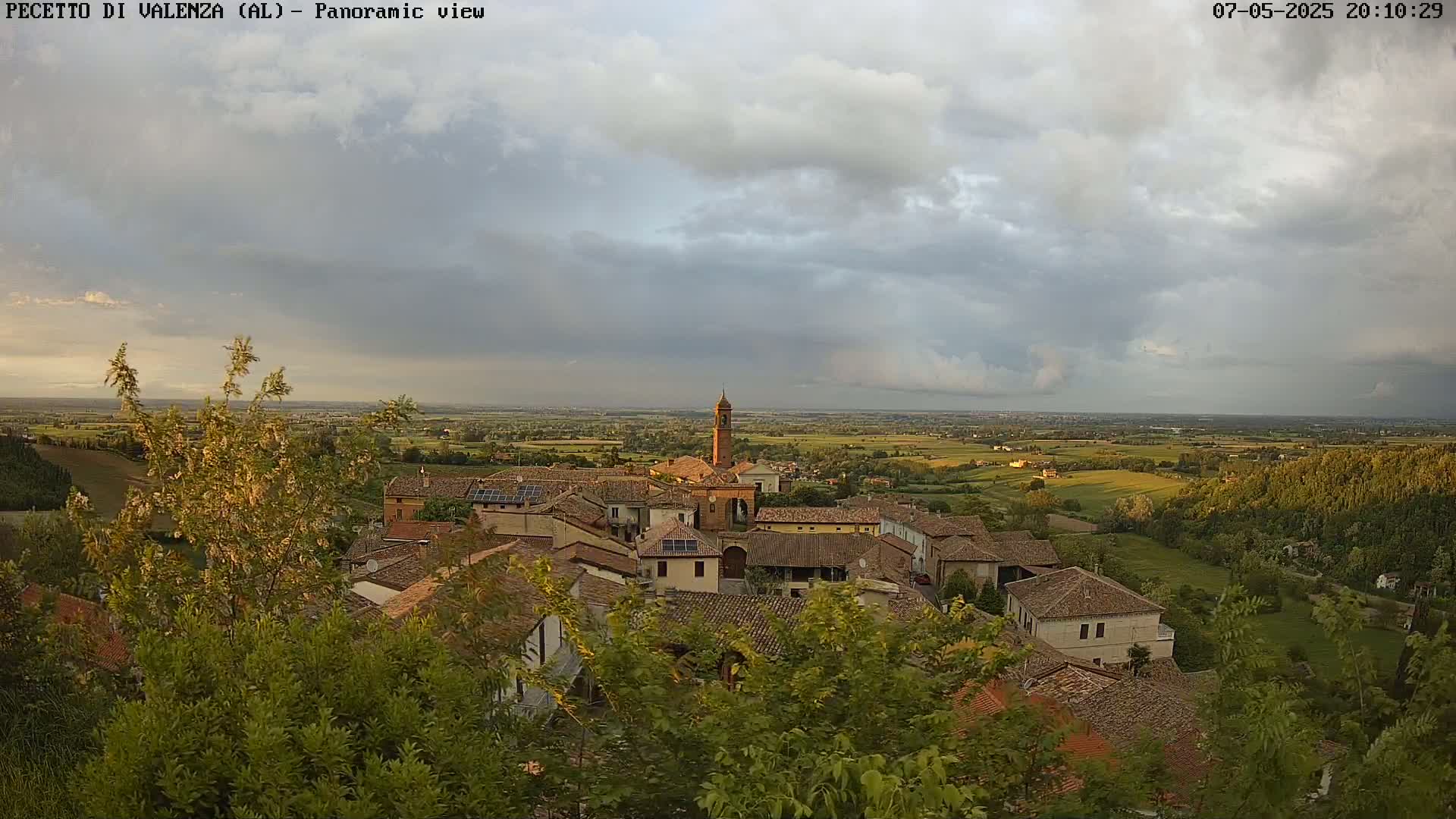 A small village with terracotta rooftops and a tall bell tower is nestled amongst green trees and rolling hills under a partly cloudy sky.
