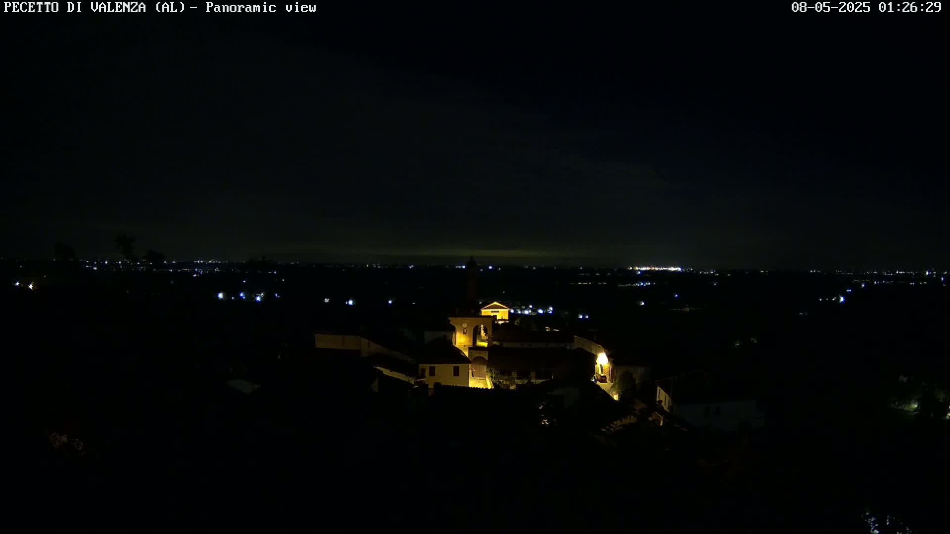 A nighttime panoramic view shows a small, illuminated village nestled in a dark landscape, with distant lights twinkling on the horizon under a clear, dark sky.