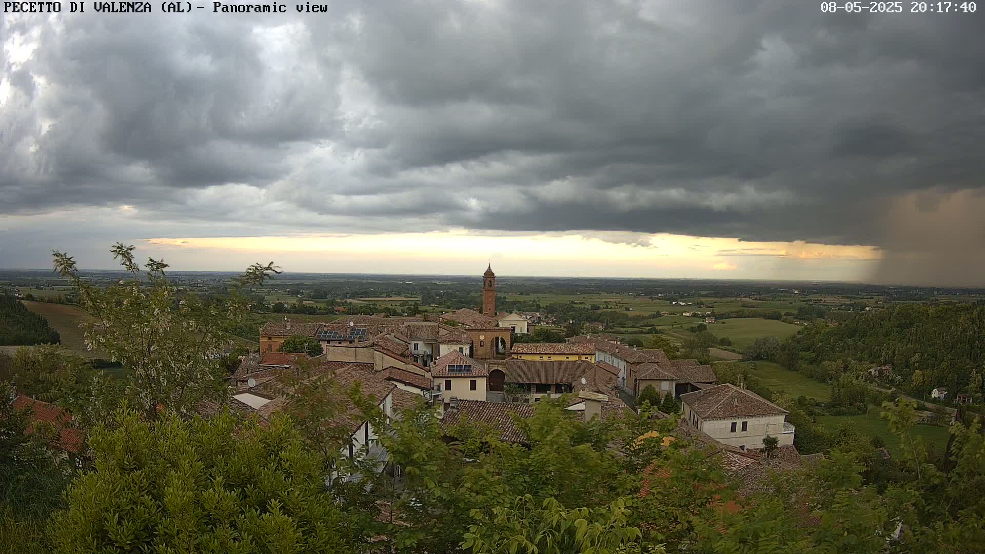 A small village with red-tiled roofs nestled in a green valley under a stormy, gray sky with a sliver of sunset visible in the distance.