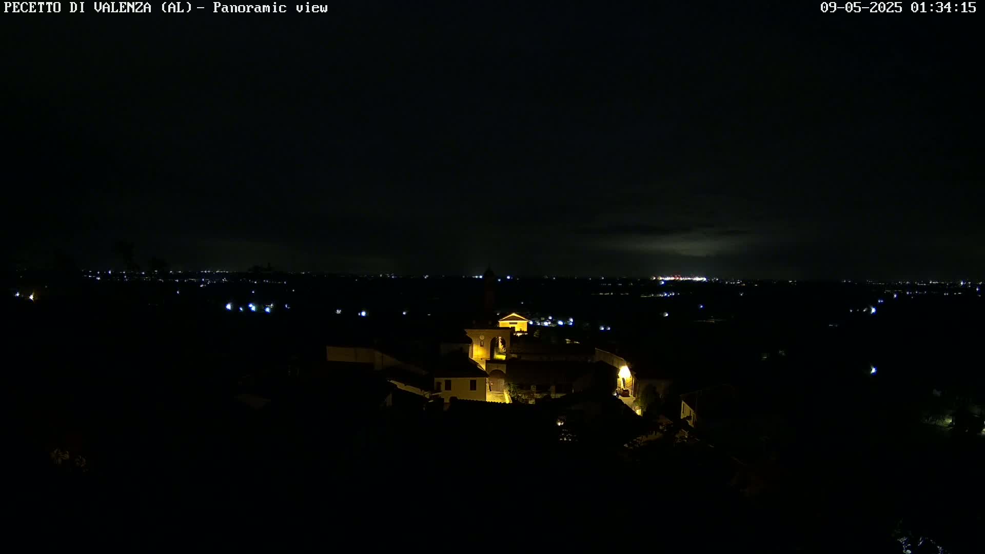 A nighttime panoramic view shows a small, illuminated town nestled in a dark landscape, with faintly visible lights in the distance under a mostly clear sky.