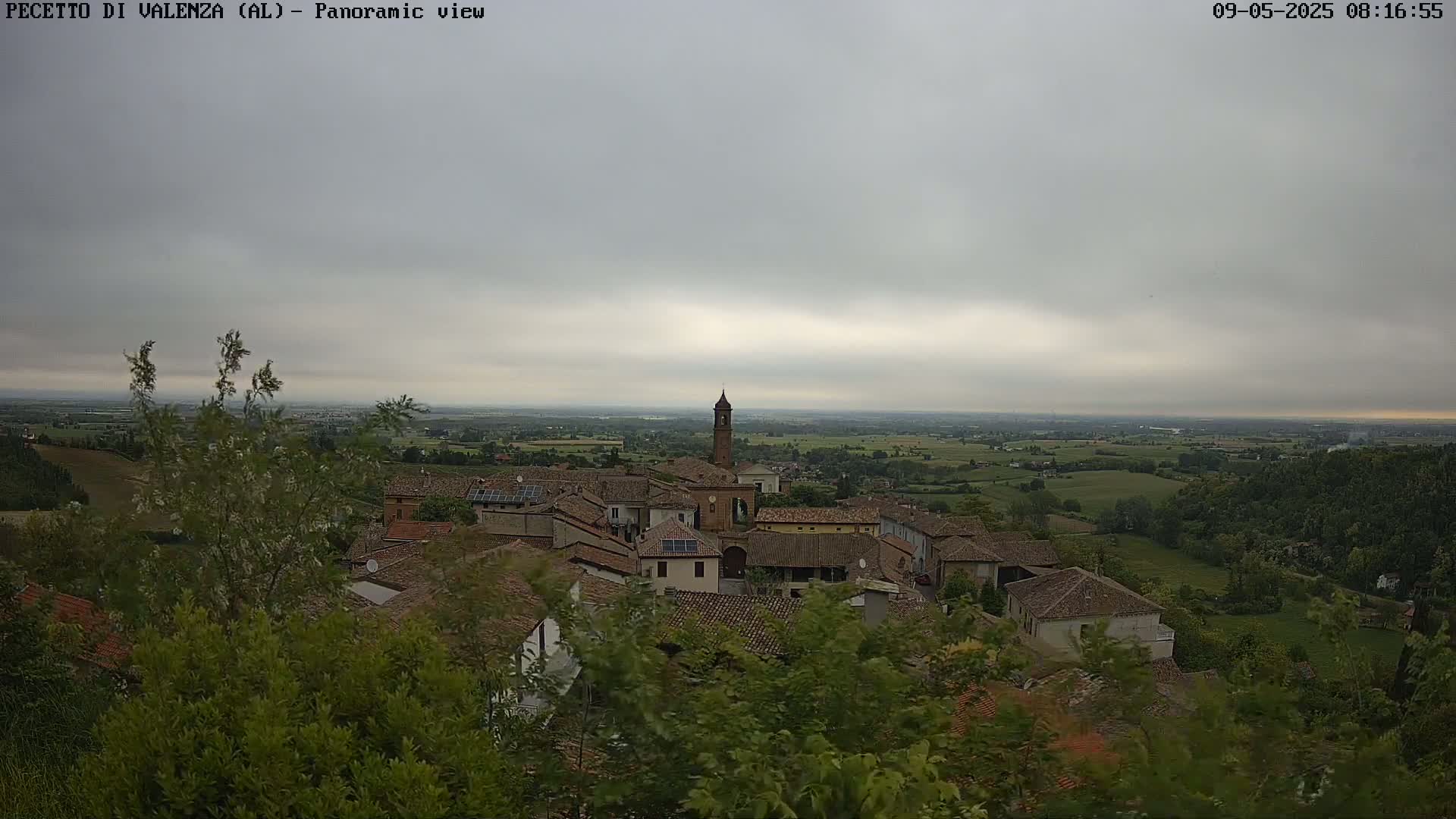 An overcast sky hangs over a small village nestled amongst green hills and fields, viewed from a slightly elevated perspective.
