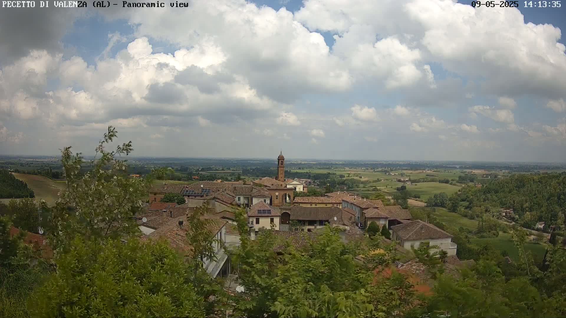 A partly cloudy sky overlooks a small village nestled in a verdant valley, with rolling hills and farmland extending to the horizon.