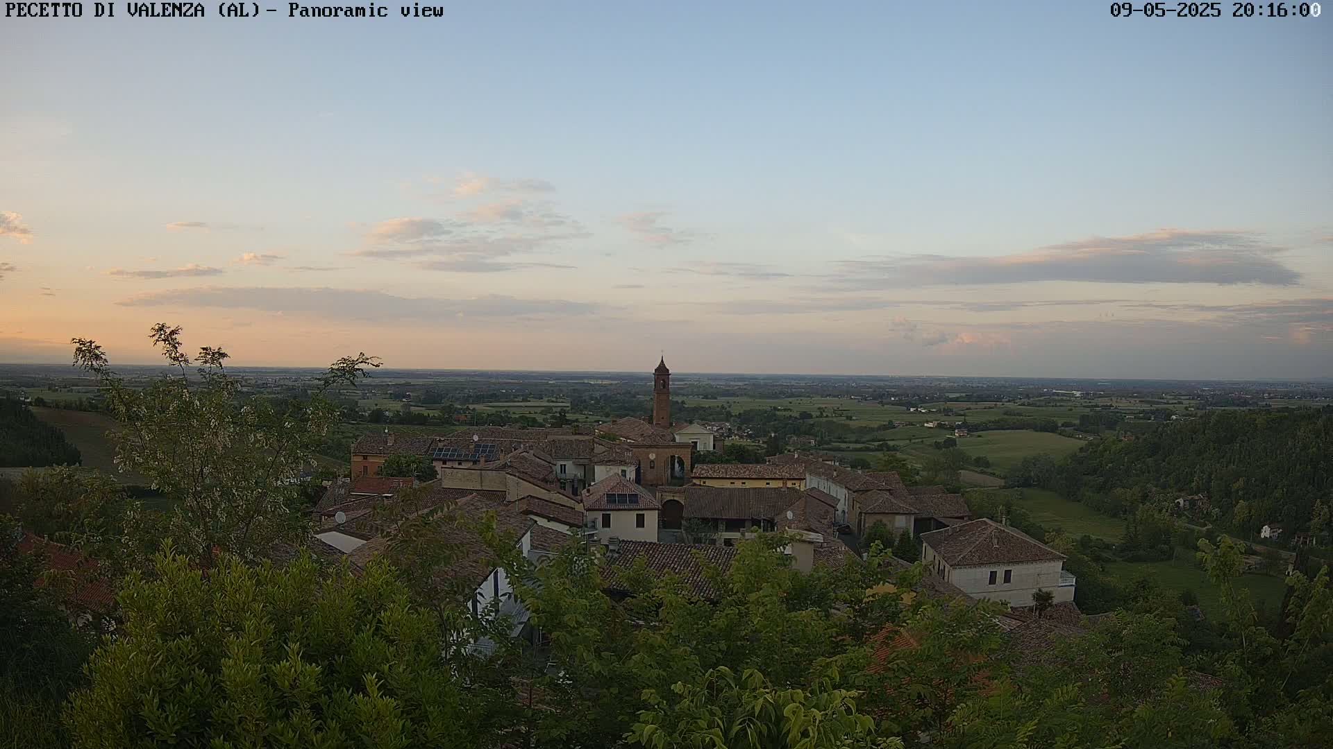 A small village with terracotta rooftops and a tall bell tower is nestled among green trees and overlooks a vast, flat landscape under a pale, dusky sky.