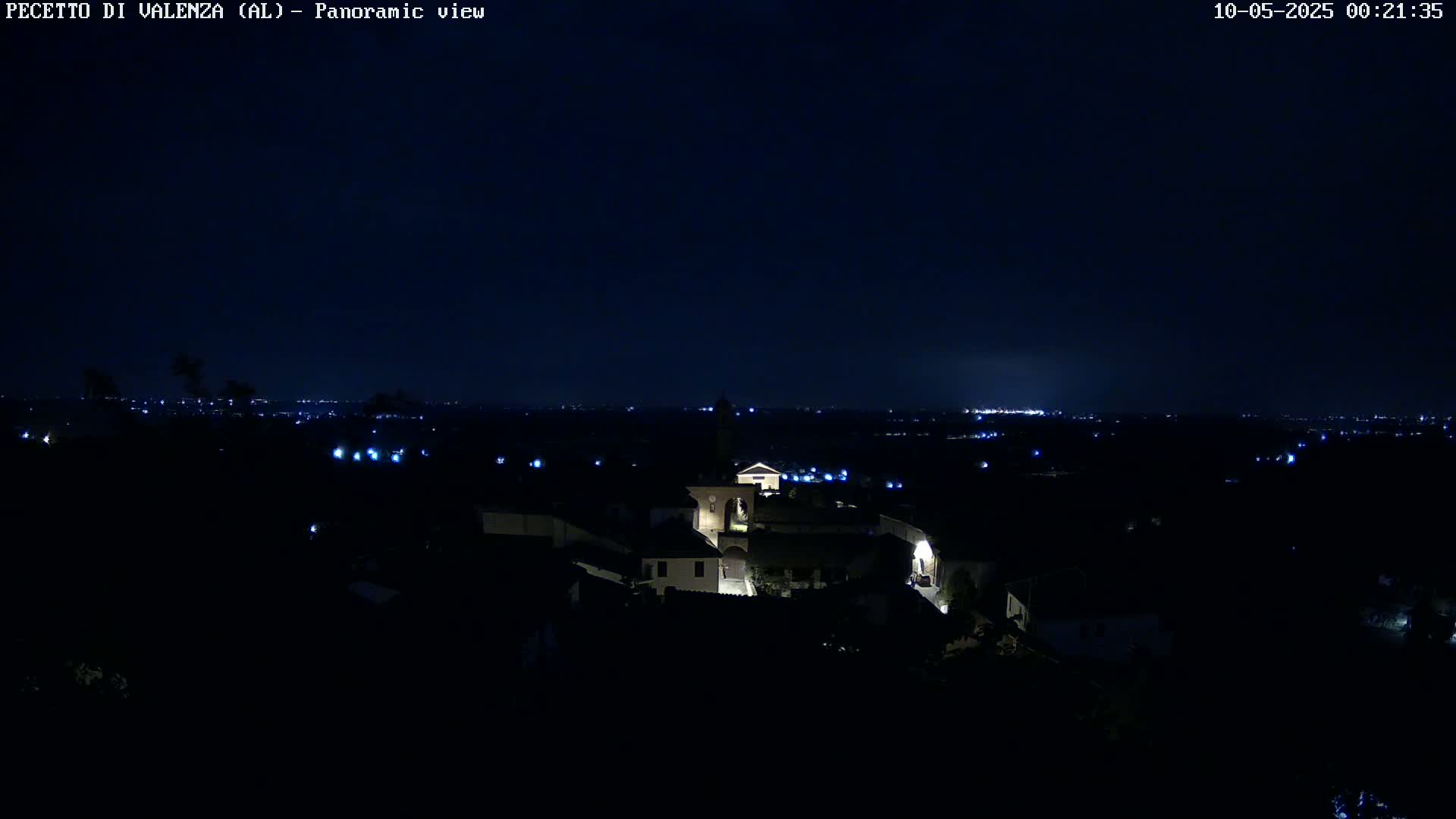 A nighttime panoramic view shows a small, illuminated town nestled in a dark landscape under a clear, dark sky.