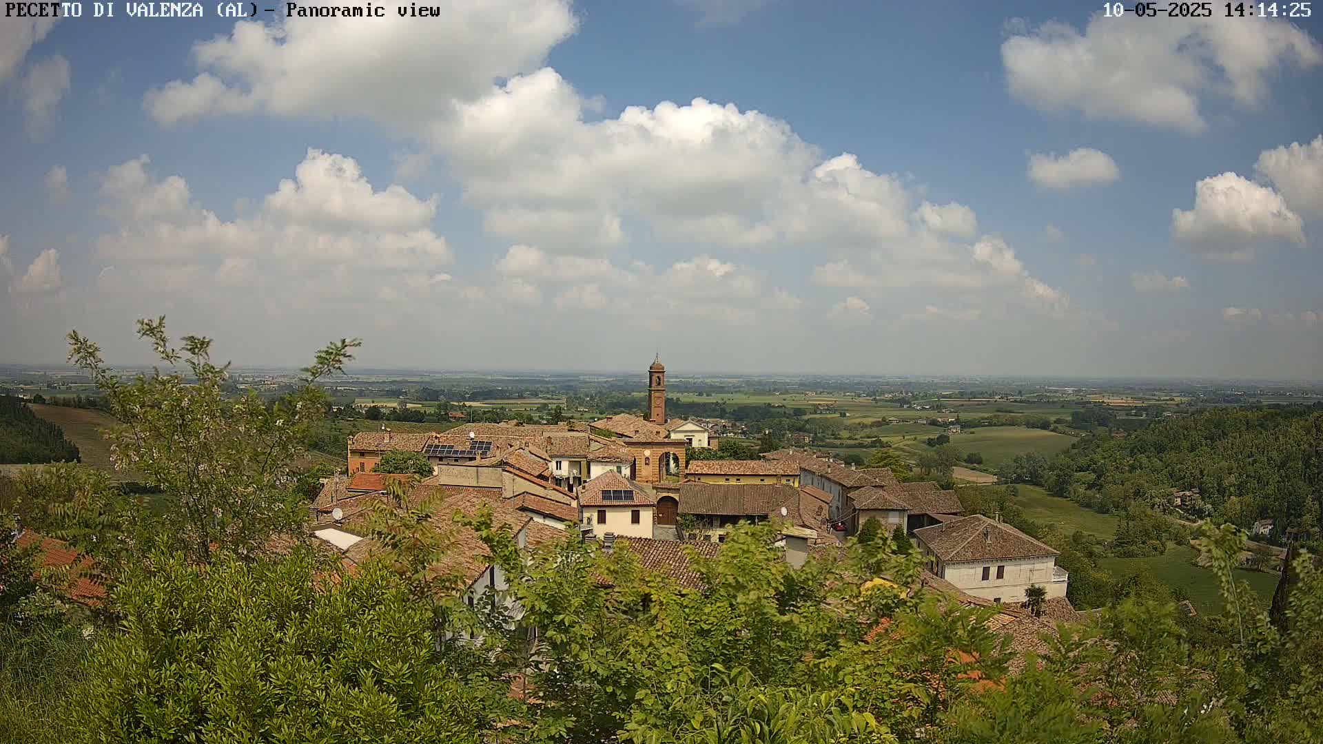 A partly cloudy day reveals a panoramic view of a small village nestled amongst green hills and farmland.
