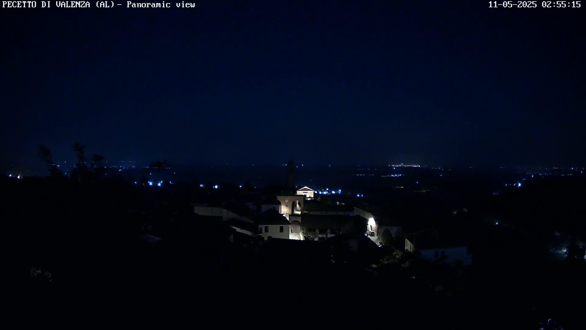 A nighttime panoramic view of a small, dimly lit village nestled in a dark landscape under a clear, starless sky.