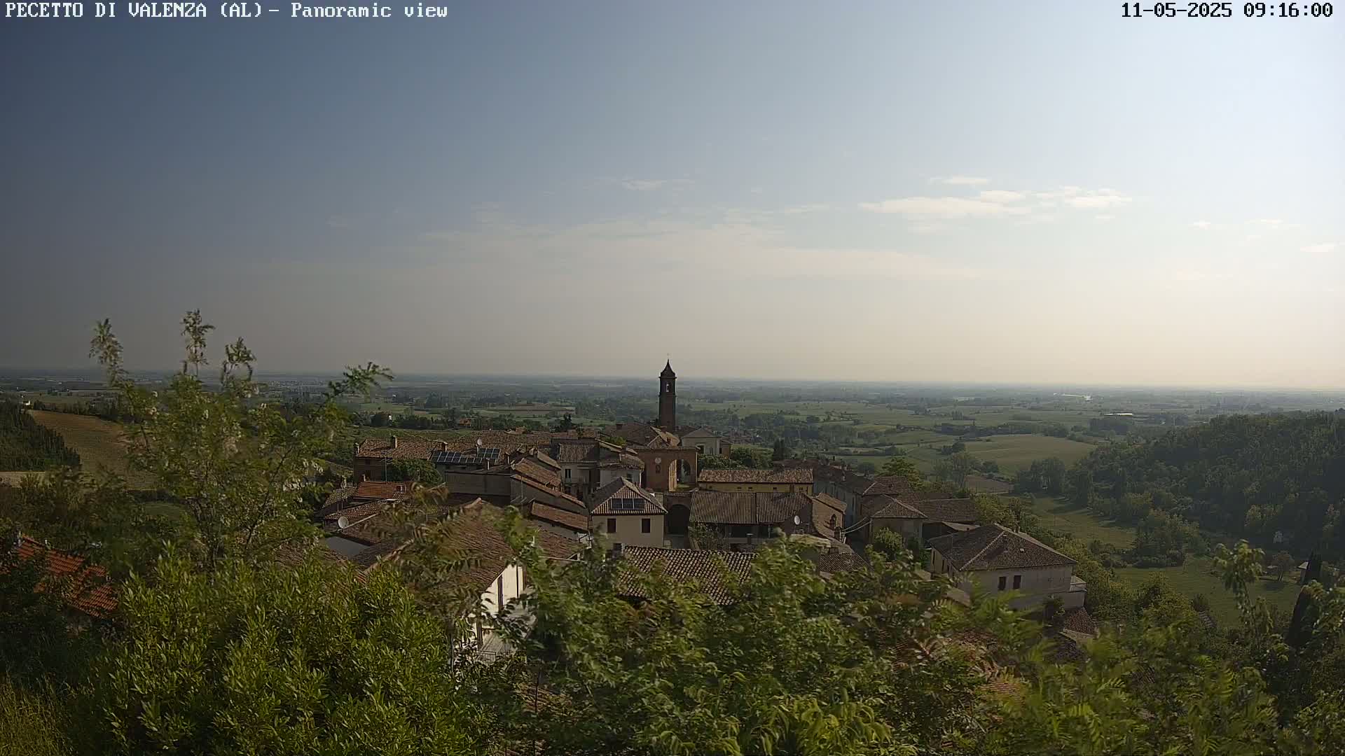 A panoramic view of a small village nestled in a verdant valley under a mostly clear, bright sky.