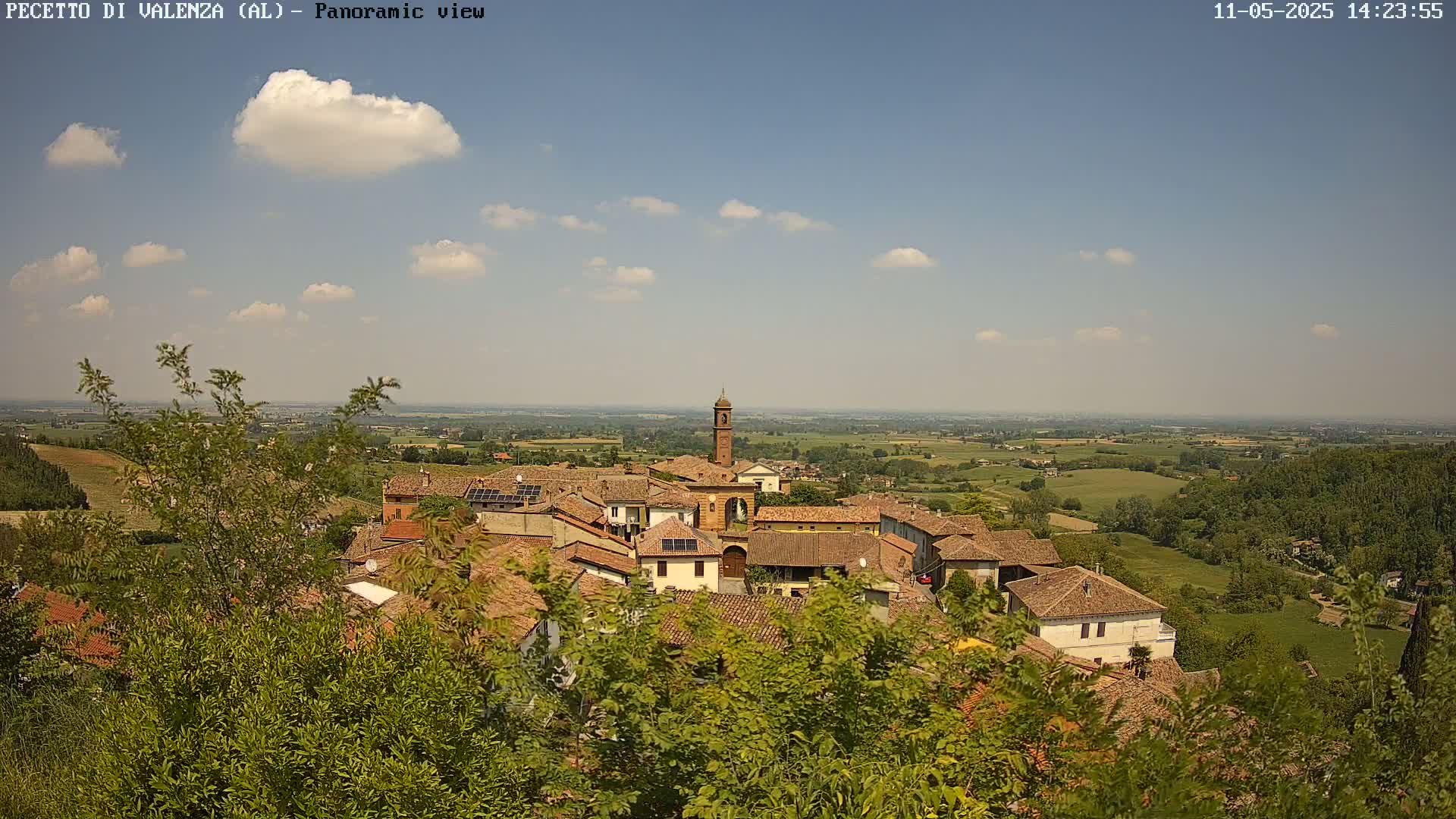 A partly cloudy, sunny day overlooks a small village of red-tiled buildings nestled in a green valley, with a church steeple at its center.