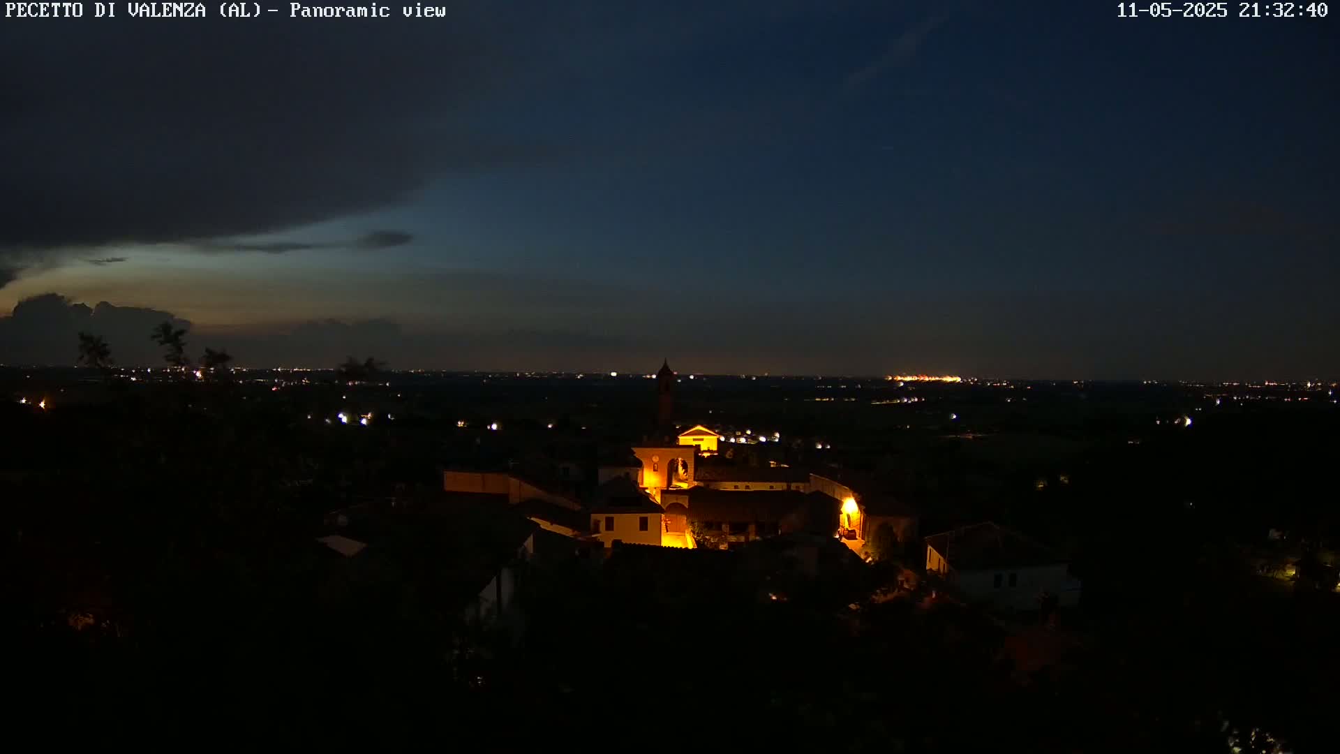 A small, illuminated village sits on a hill under a mostly dark night sky with a few clouds, overlooking a dimly lit plain.
