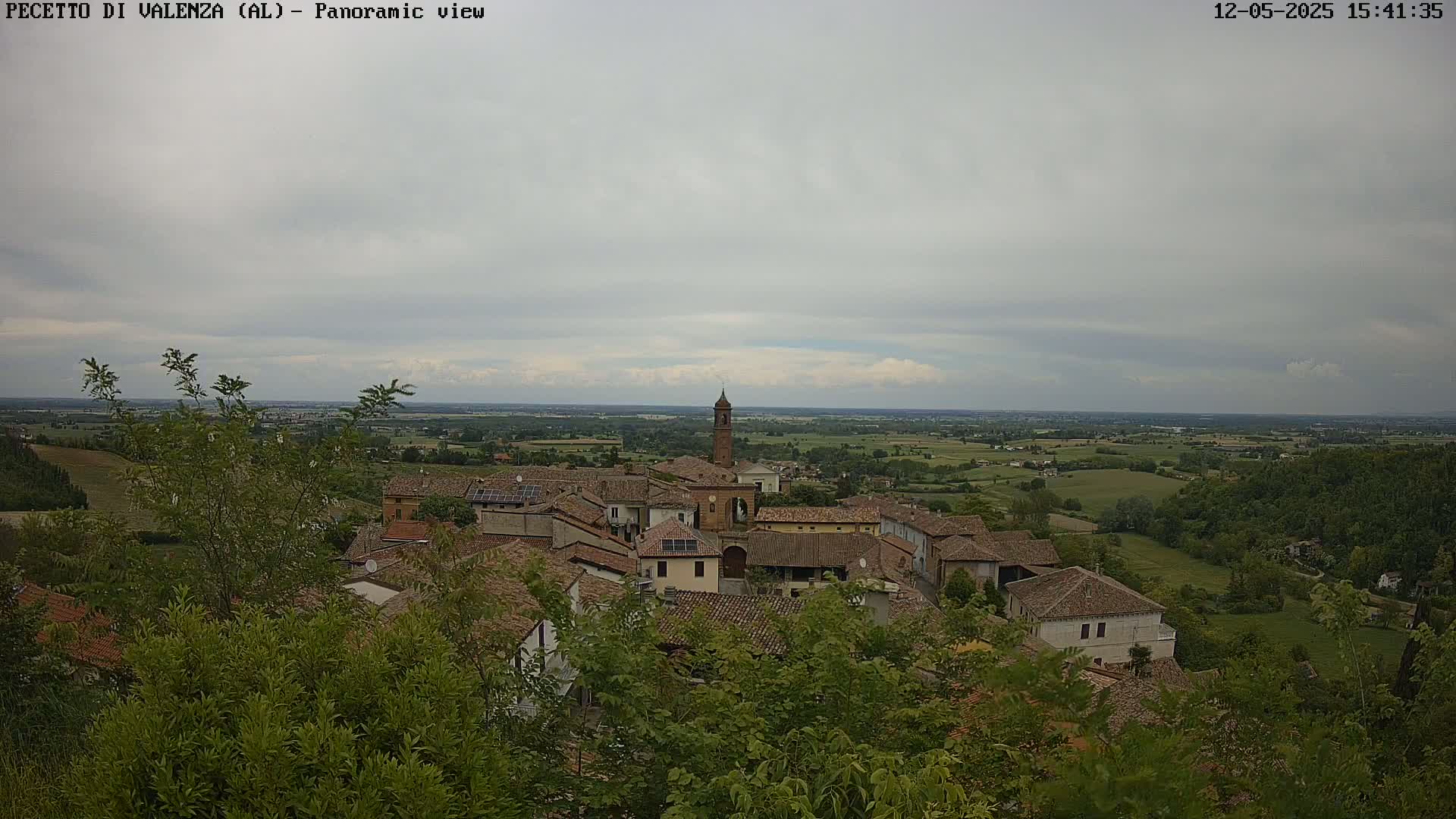 A small town with red-tiled roofs and a tall bell tower is nestled in a verdant valley under a mostly cloudy sky.