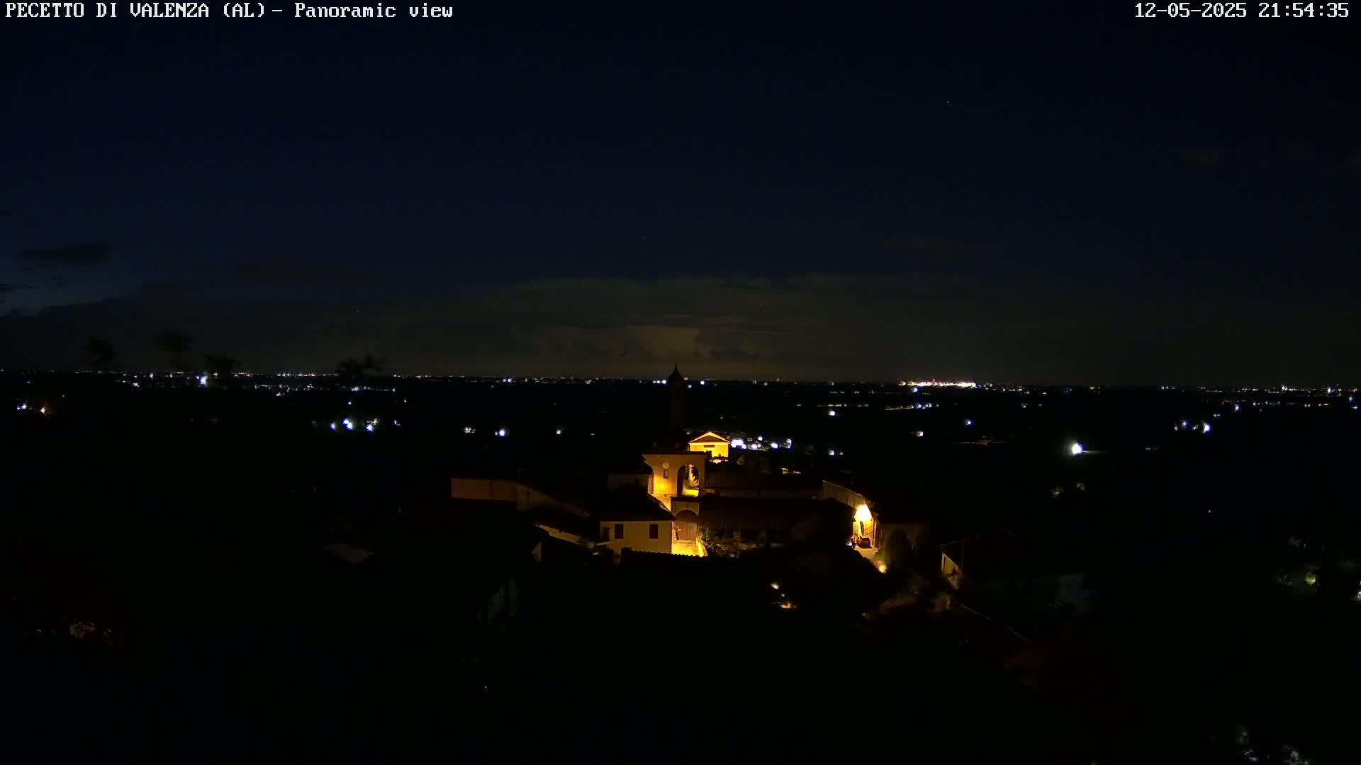 A nighttime panoramic view shows a small, illuminated village nestled in a dark landscape under a mostly clear, starry sky.
