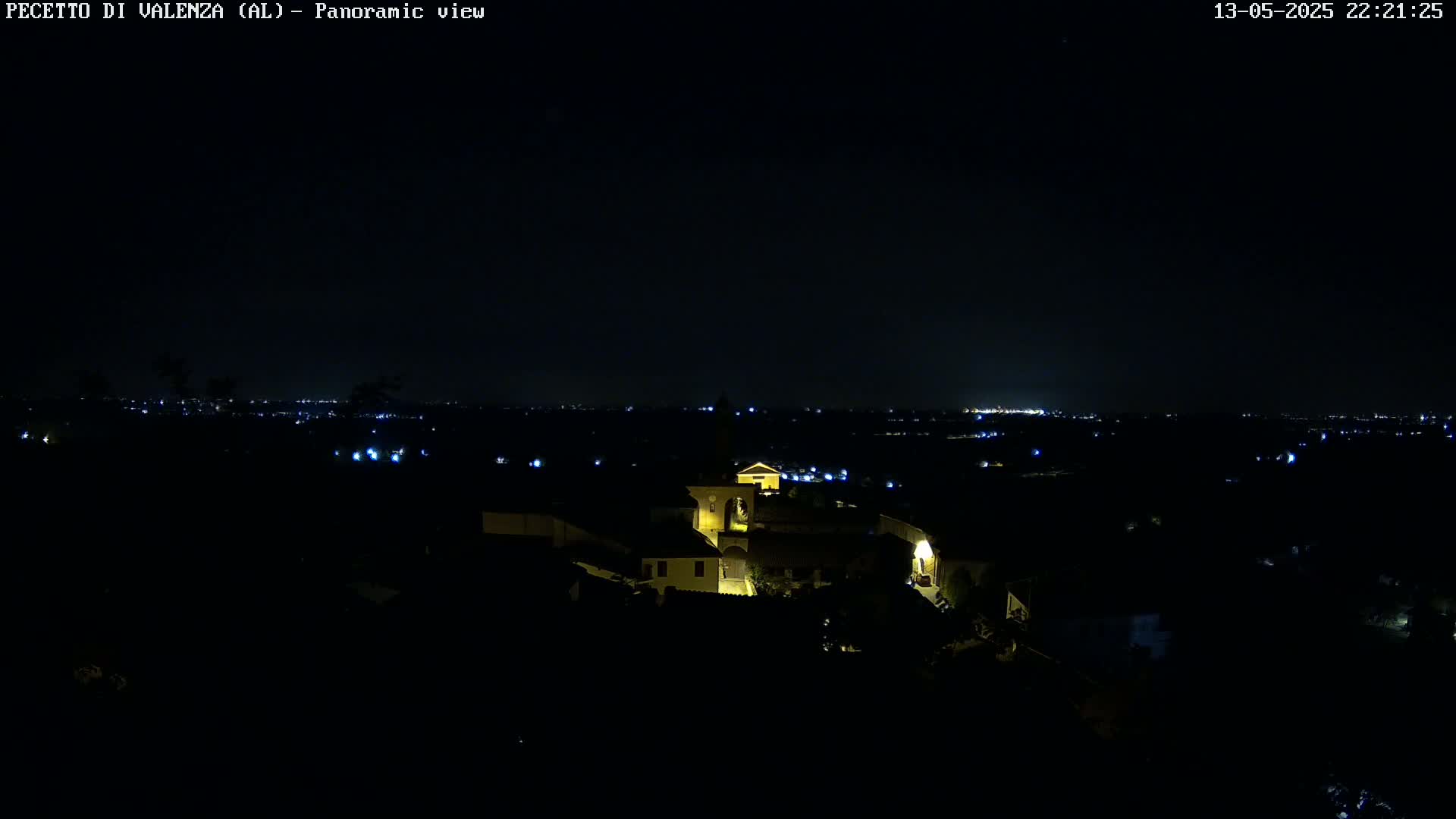 A nighttime panoramic view shows a small, illuminated village nestled in a dark landscape, with a distant city skyline faintly visible under a clear, dark sky.