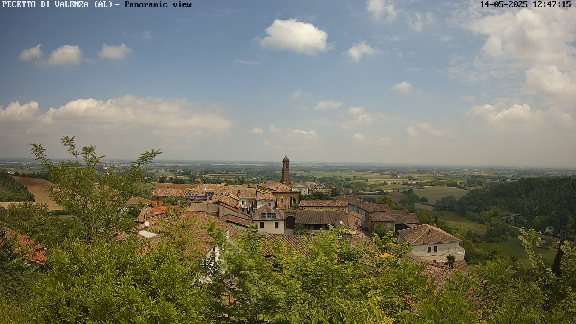 A partly cloudy sky overlooks a small town of terracotta-roofed buildings nestled in a verdant landscape.
