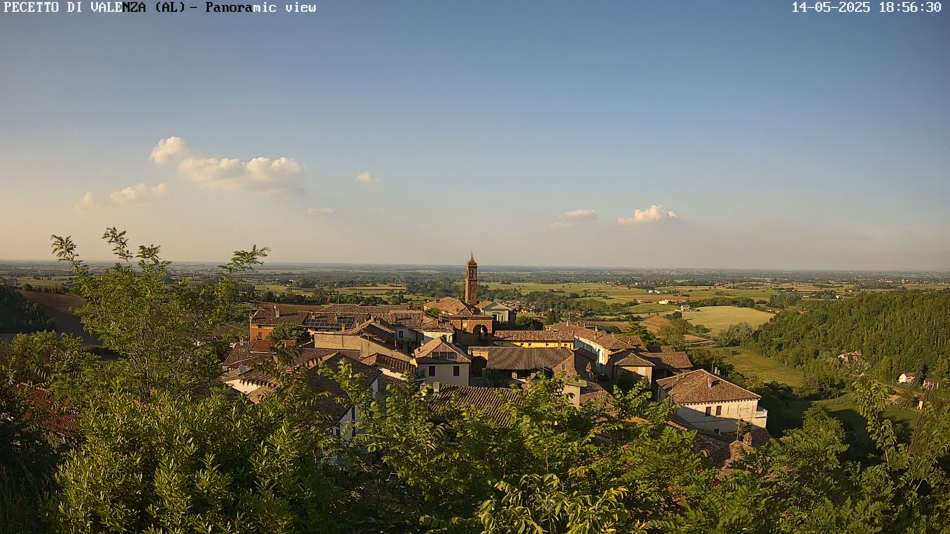 A partly cloudy, sunny day overlooks a small village nestled among green trees and rolling hills.