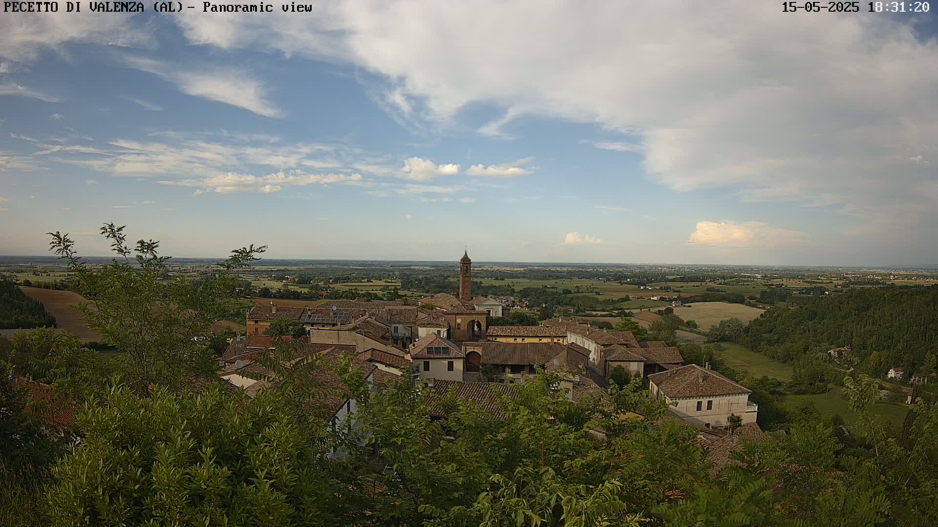 A partly cloudy sky overlooks a village of brown-roofed buildings nestled among green trees and fields, extending to a distant horizon.