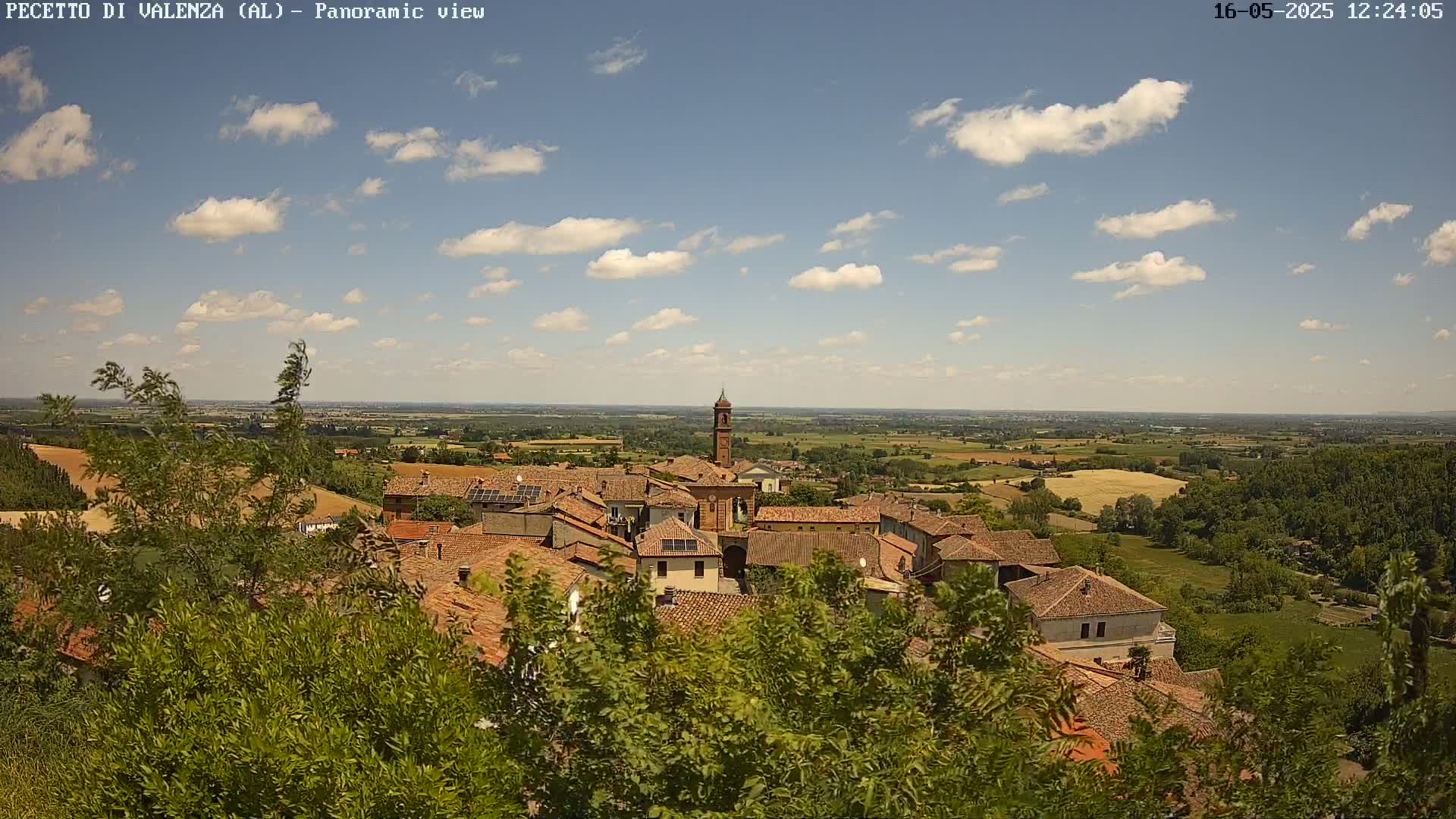 A small town with red-tiled roofs nestles amongst green vegetation, overlooking a vast, sunny landscape under a partly cloudy sky.