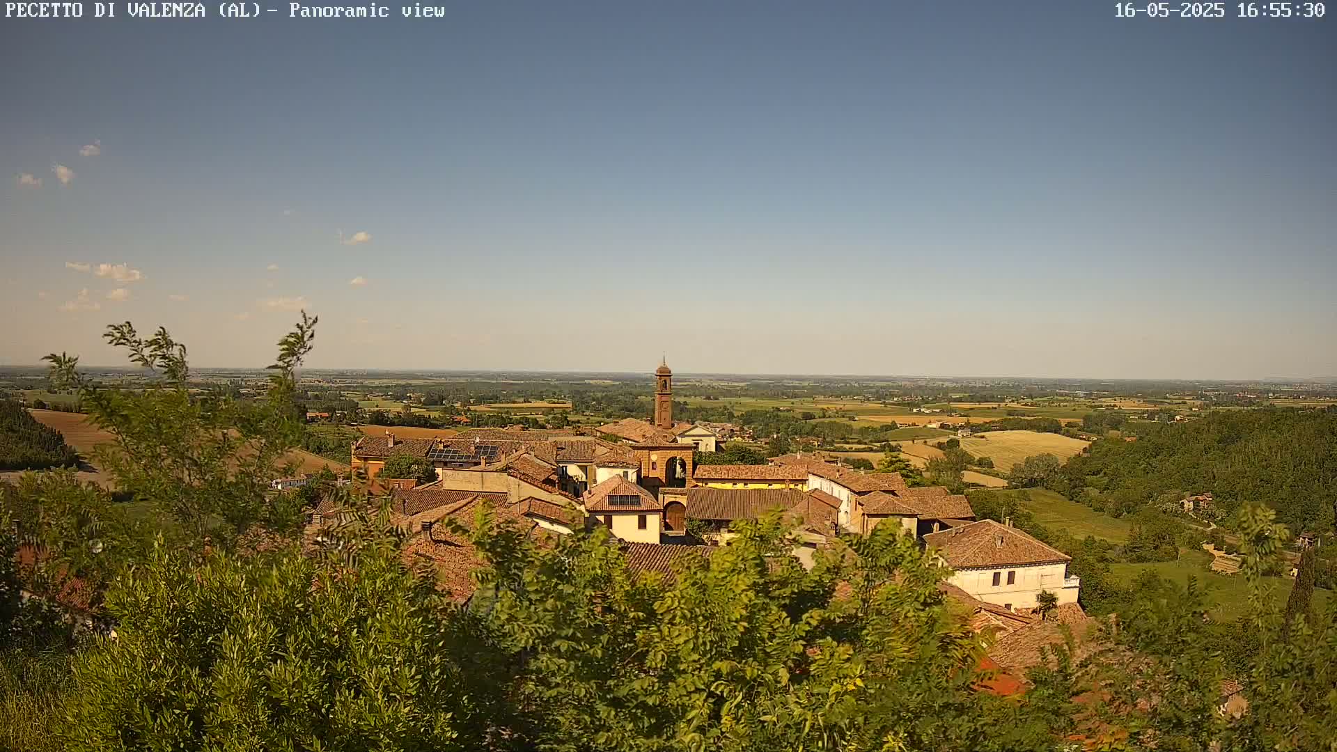 A panoramic view of a small village nestled amongst green hills and fields under a mostly clear, light blue sky.