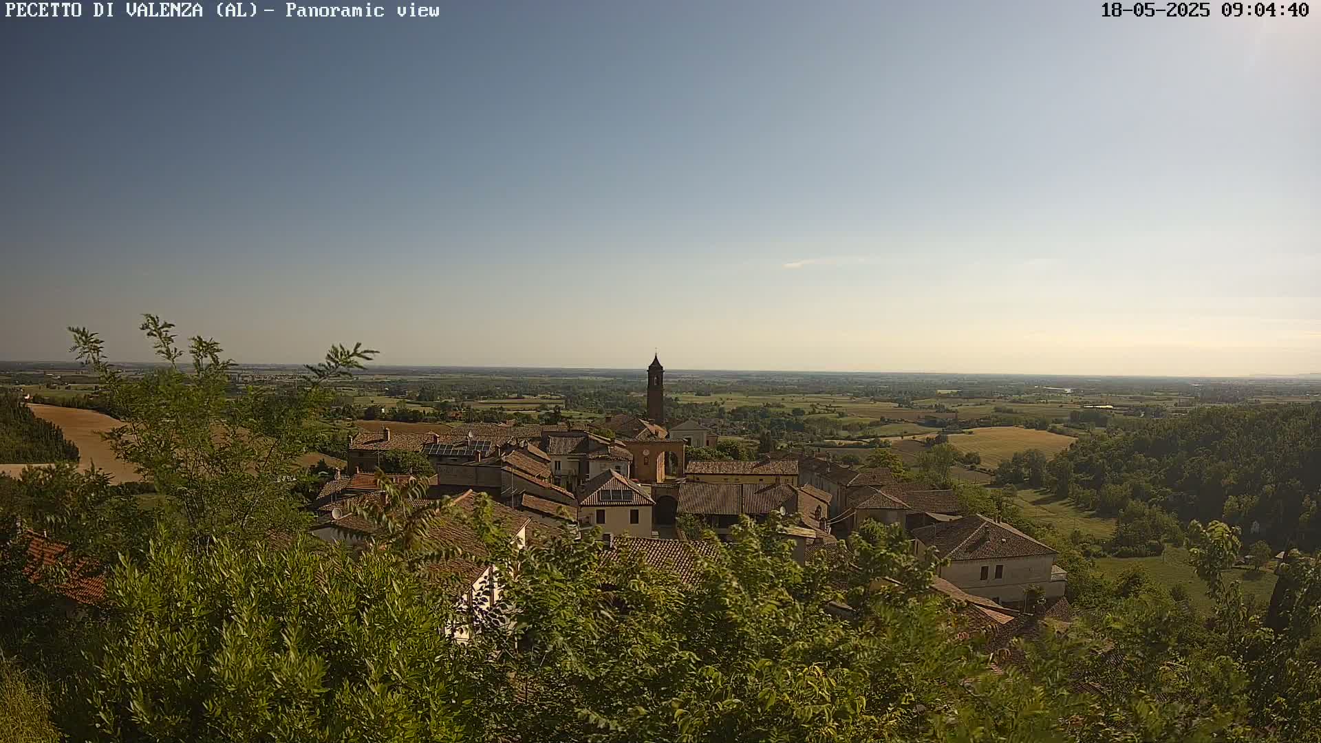 A small town with red-tiled roofs nestles in a valley under a clear, bright sky, surrounded by lush green vegetation and farmland extending to the horizon.