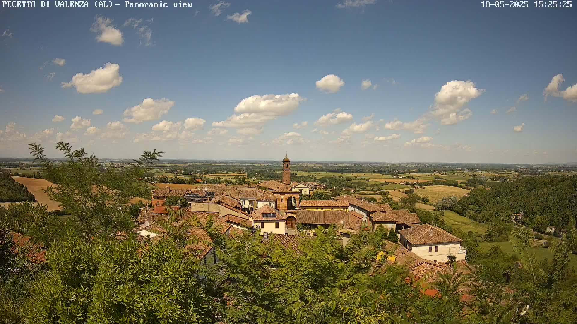 A partly cloudy sunny day overlooks a small town with terracotta rooftops and a tall bell tower, nestled in a verdant landscape of fields and forests.