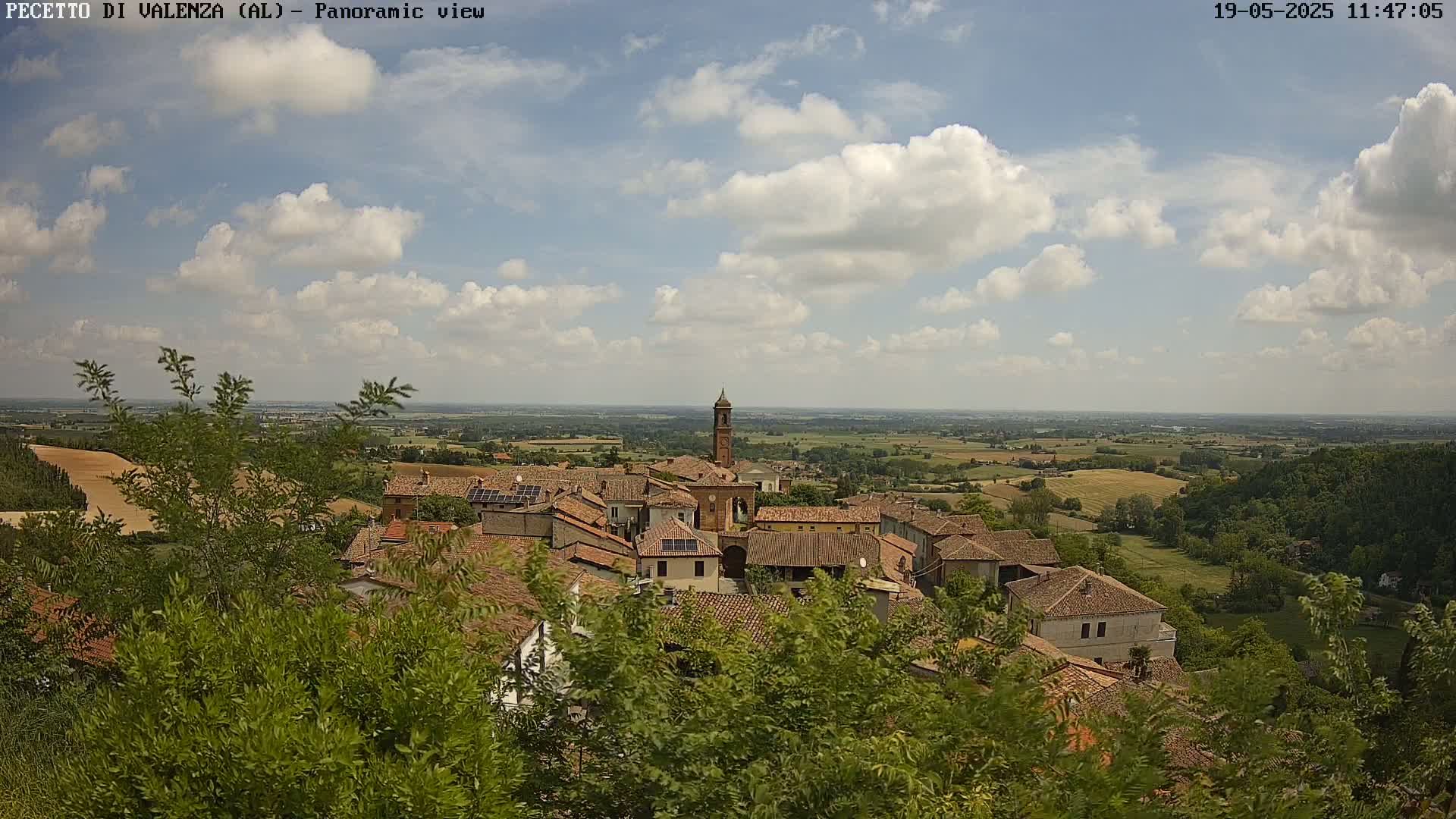 A partly cloudy sky overlooks a small village nestled in a verdant valley, with fields stretching to the horizon.