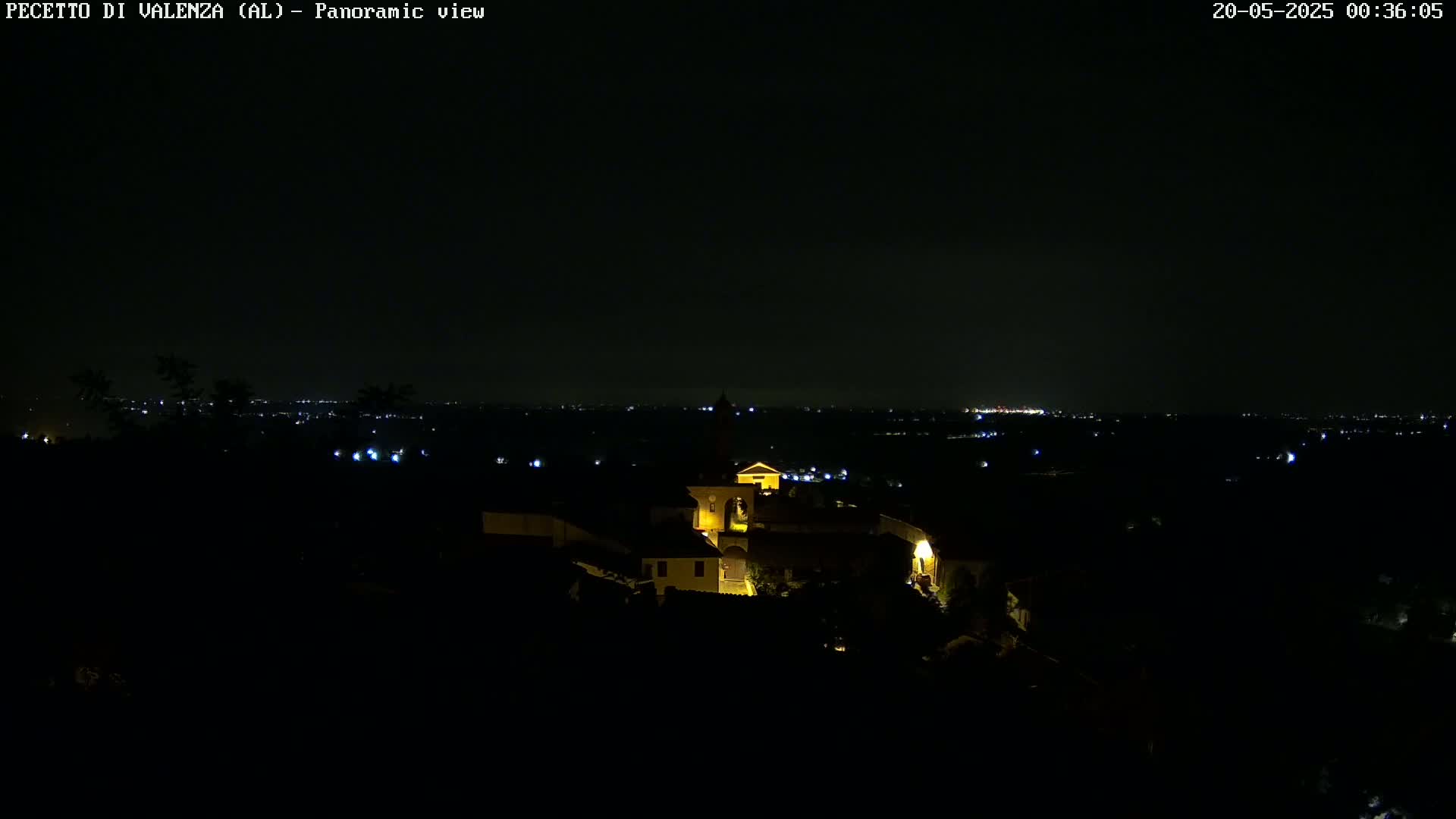 A nighttime panoramic view shows a small, illuminated village nestled in a dark landscape, with a distant city skyline faintly visible on the horizon under a clear, dark sky.