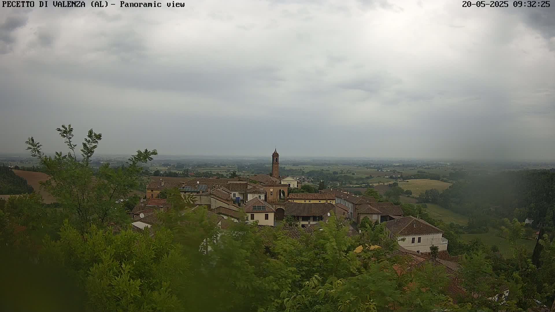 An overcast day reveals a panoramic view of a small village nestled among green hills and farmland, with light rain falling.