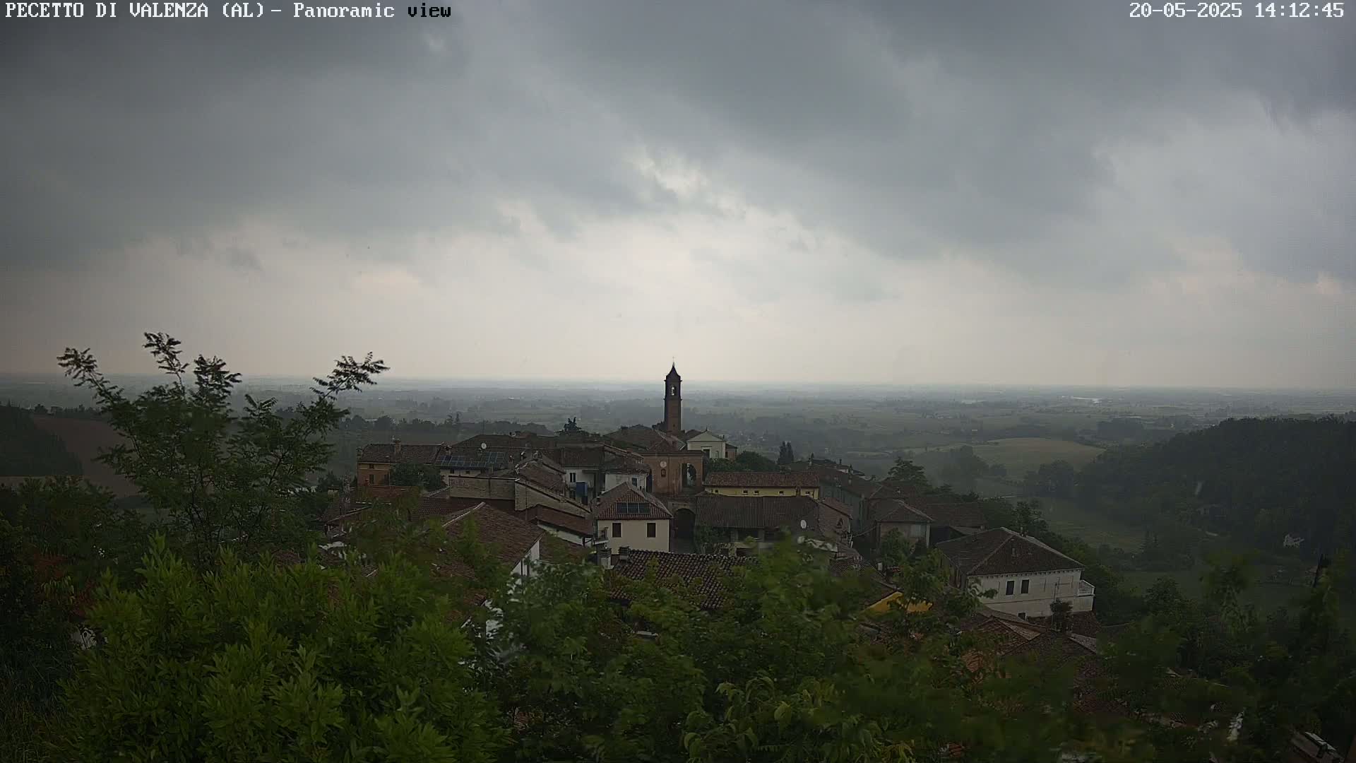 A small village with a bell tower is nestled among green foliage under a cloudy, overcast sky.