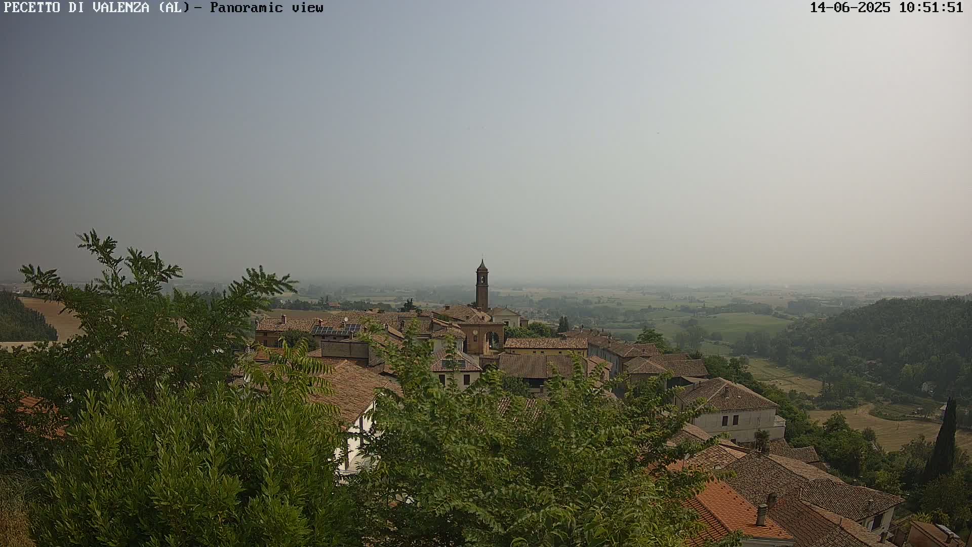 A hazy, panoramic view shows a village of brown-roofed buildings nestled among green trees, with a tall bell tower visible in the center, overlooking a wide, flat expanse of farmland.