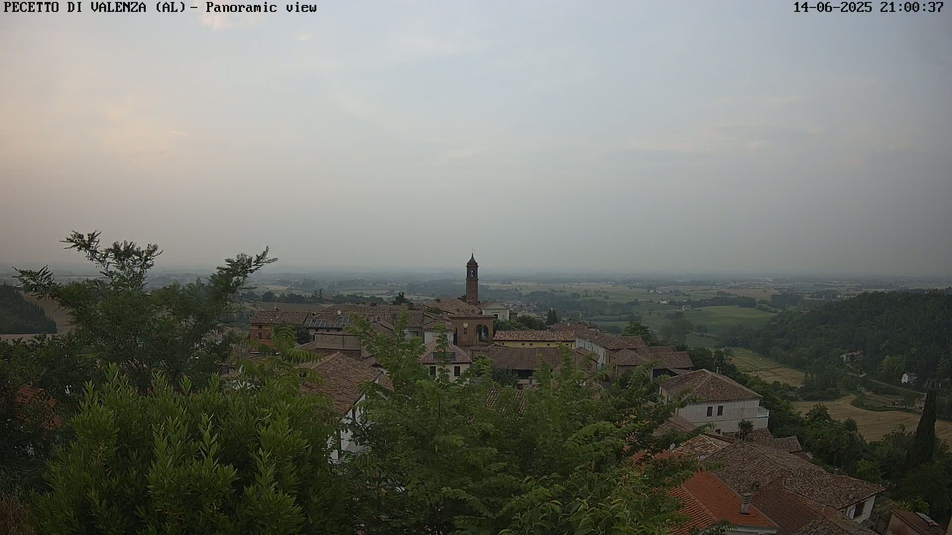 A hazy, sunny day overlooks a small village of terracotta-roofed buildings nestled in a verdant valley.