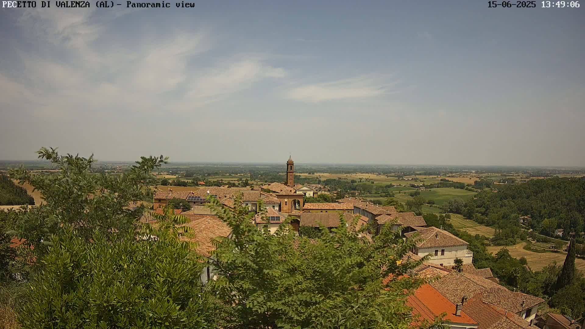 A mostly sunny day reveals a panoramic view of a small town nestled in a verdant valley, with terracotta rooftops and a prominent bell tower visible amongst the buildings.