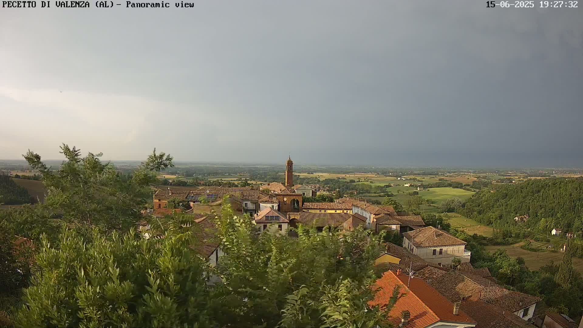 An elevated view of a small town with terracotta roofs, surrounded by green fields and trees under a mostly cloudy sky.