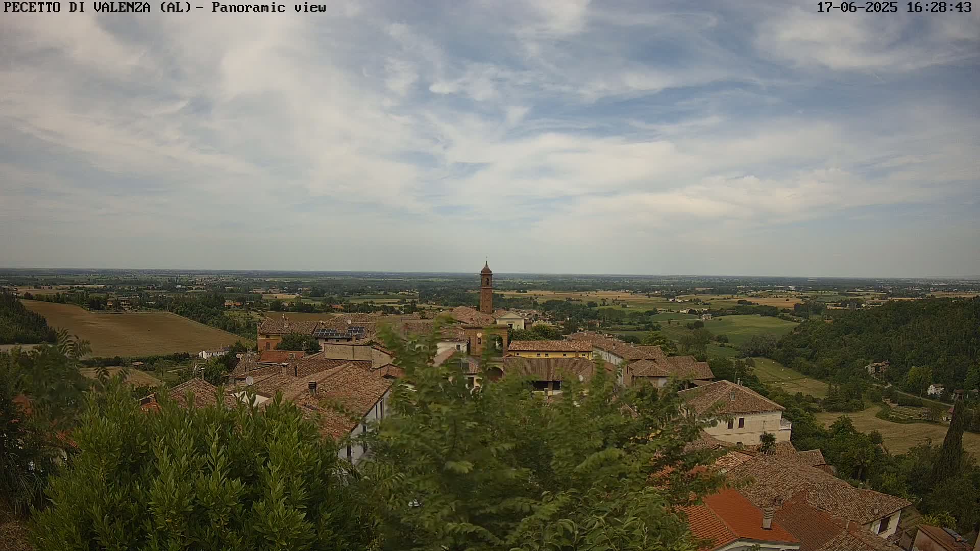 A panoramic view of a small village with terracotta rooftops nestled in a verdant landscape under a partly cloudy sky.