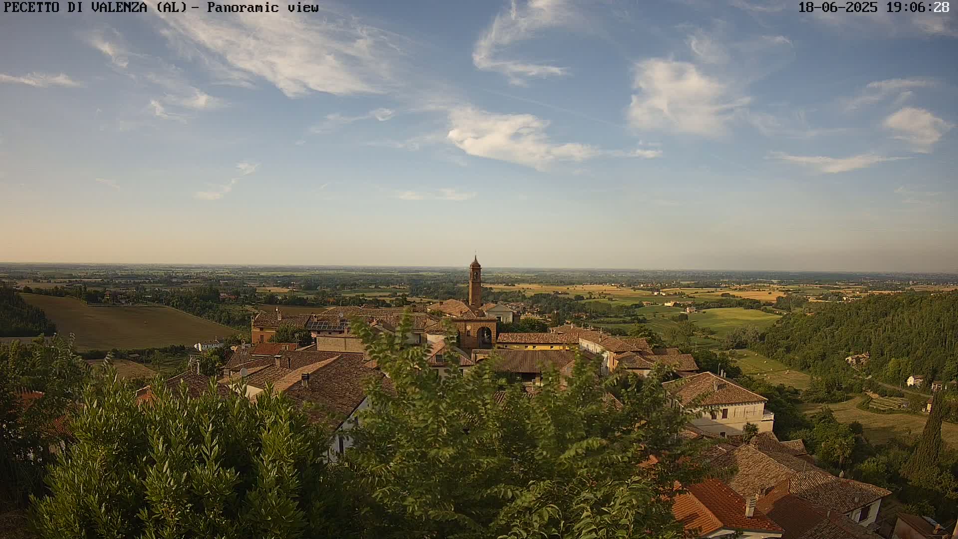 A panoramic view of a village with red-tiled roofs nestled amongst green trees and shrubs, overlooking a vast, flat landscape under a partly cloudy sky.