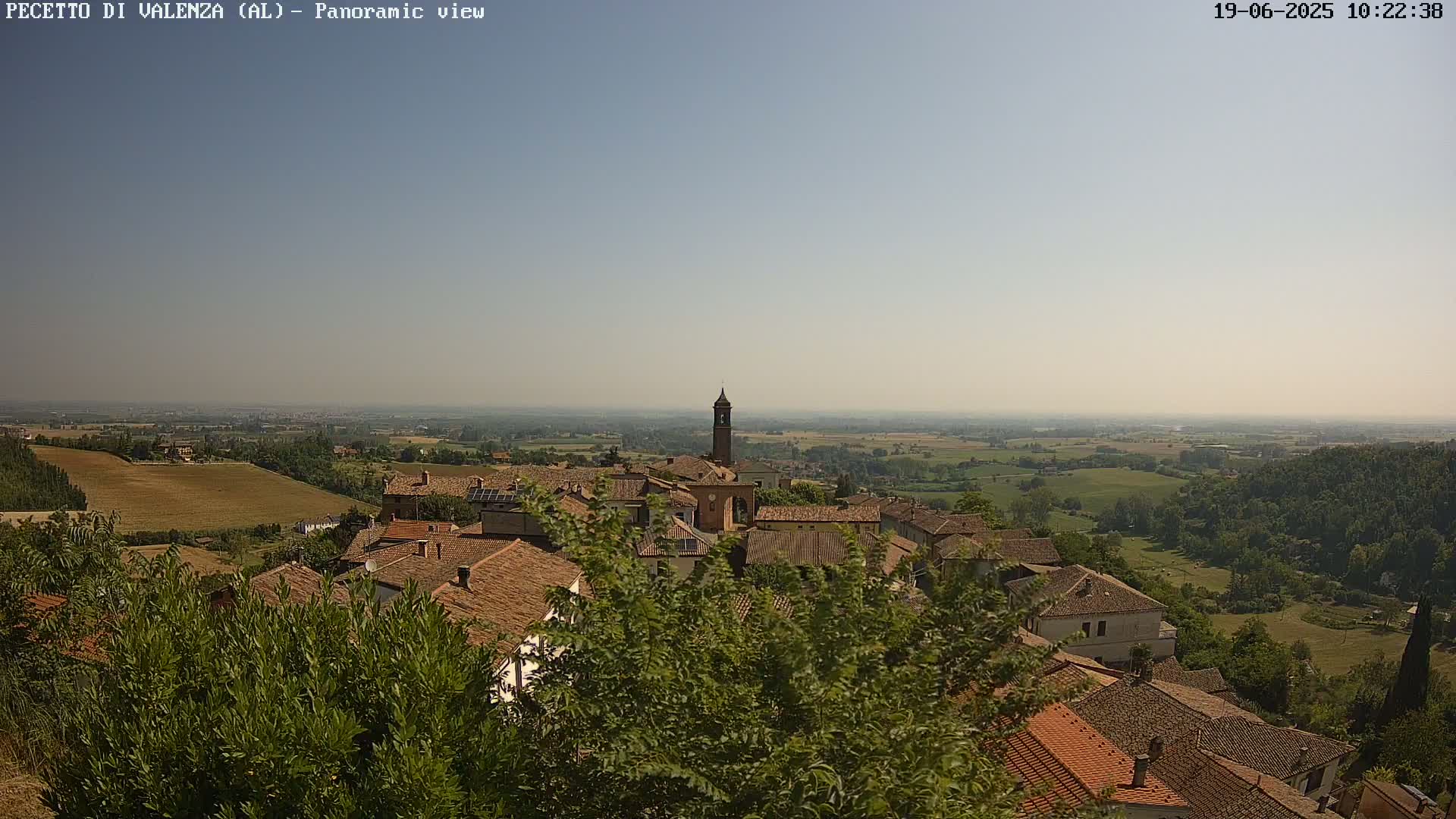 A hazy, sunny day reveals a panoramic view of a small village nestled in a hilly landscape, with terracotta rooftops and a tall bell tower prominent among the buildings.