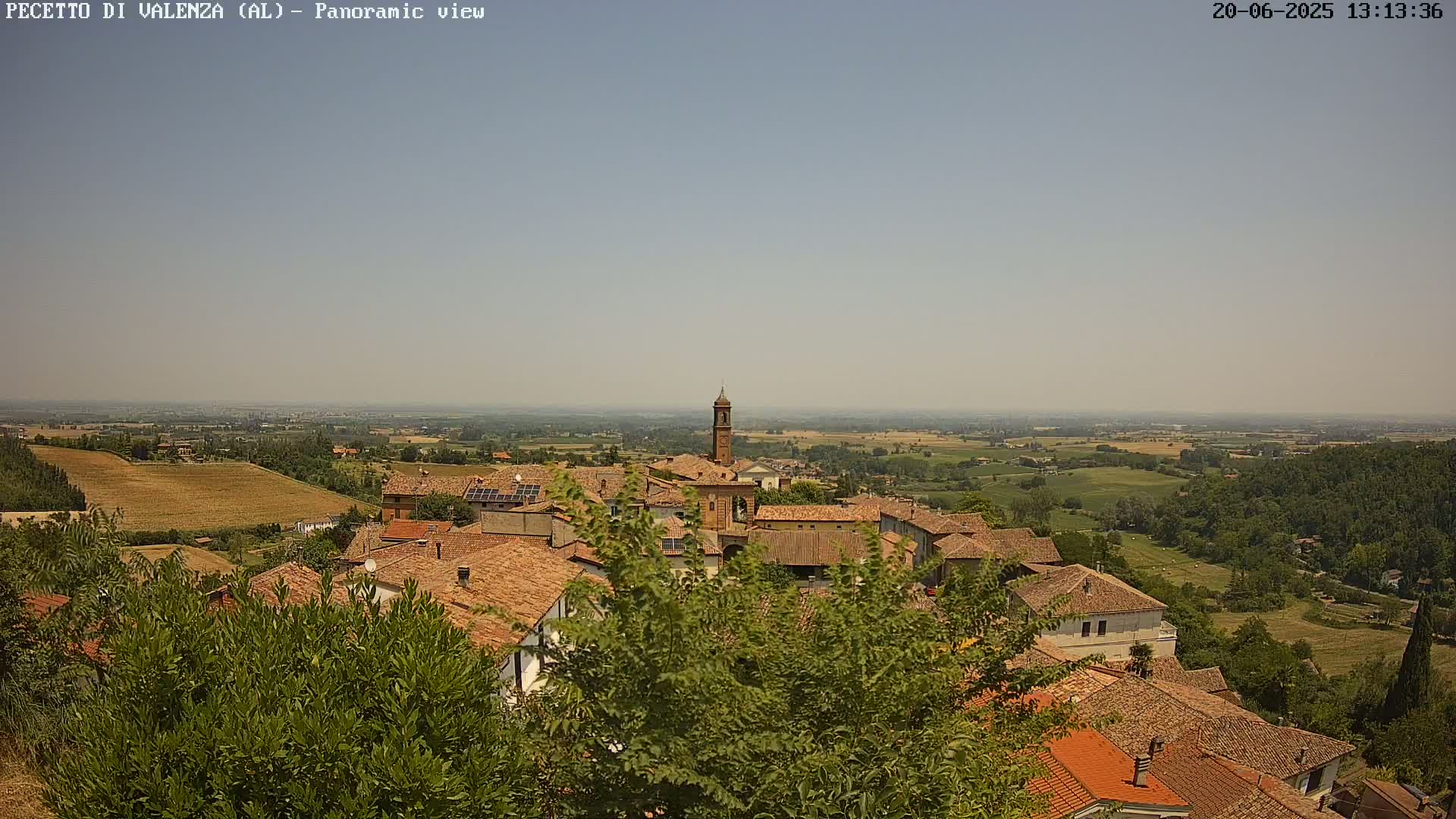 A panoramic view shows a village of brown-roofed buildings nestled among green vegetation, under a hazy, sunny sky, overlooking a vast, flat landscape.