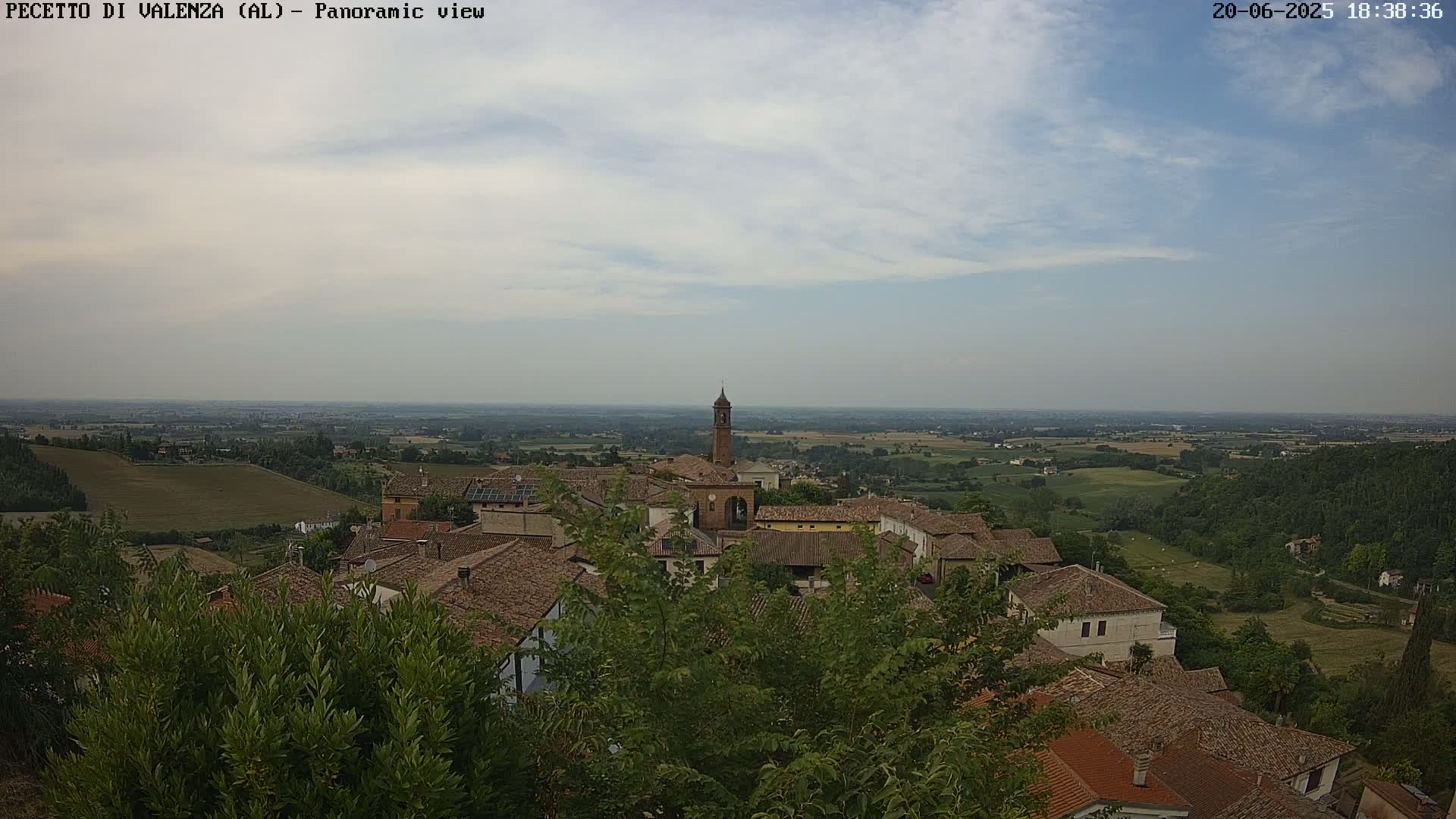 A panoramic view shows a village with terracotta roofs nestled among green foliage, overlooking a vast, flat landscape under a mostly cloudy sky.