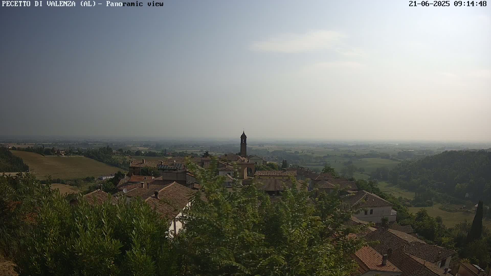 A hazy, sunny day reveals a panoramic view of a quaint village nestled among rolling green hills and farmland.