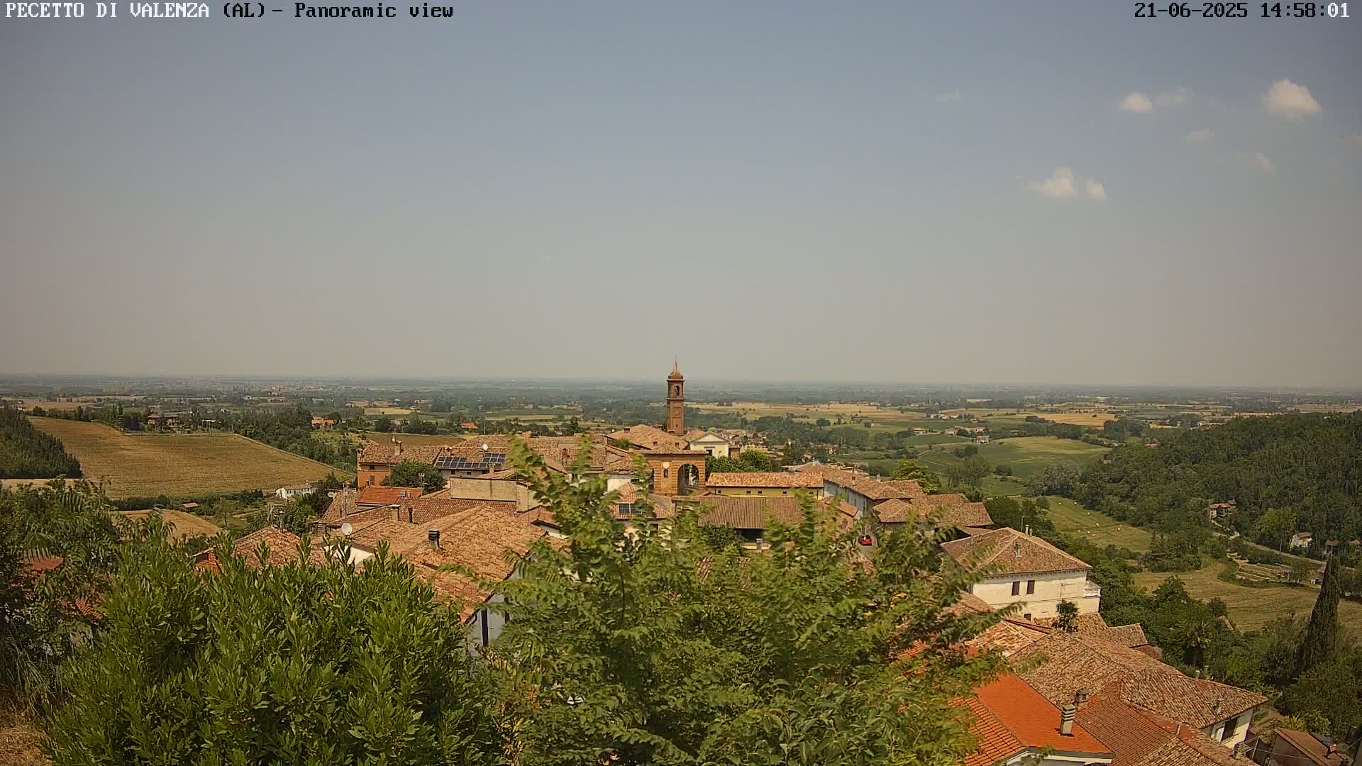 A panoramic view of a town with terracotta rooftops, a bell tower, and surrounding farmland under a mostly clear, sunny sky.