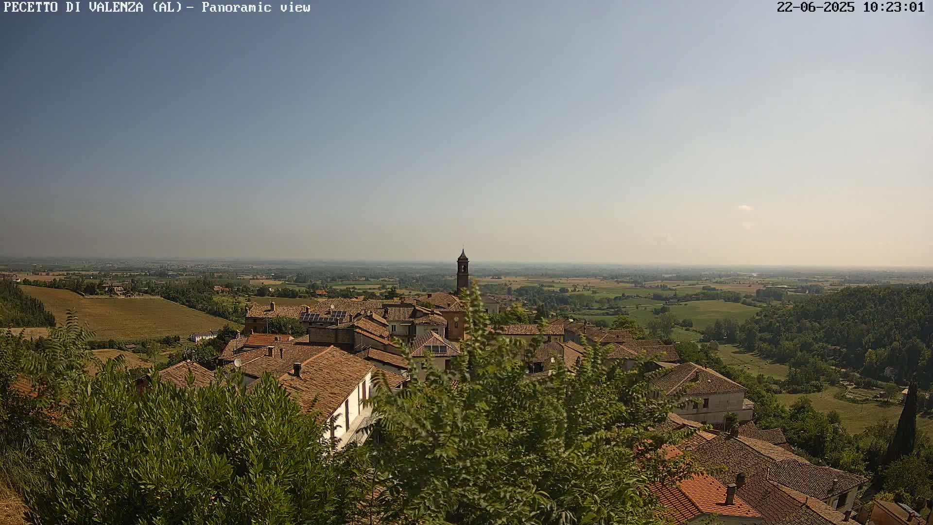 A panoramic view shows a village of buildings with red tile roofs nestled among green trees and shrubs, overlooking a vast, flat, green landscape under a clear, hazy sky.