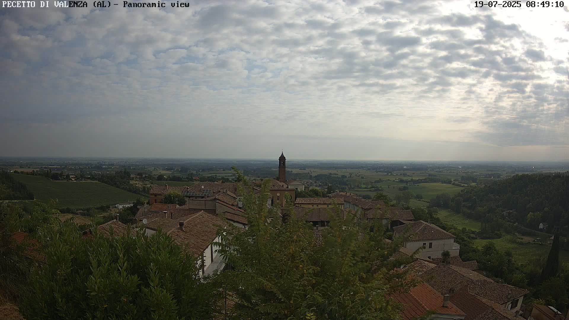 A panoramic view of a village nestled in a hilly landscape under a cloudy sky, showing red-tiled roofs and a tall bell tower.