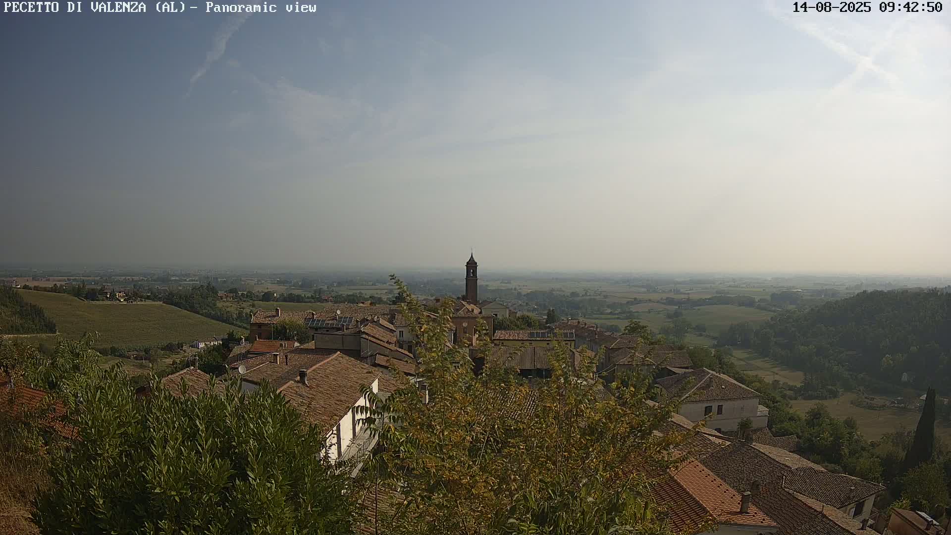 A hazy, sunny day reveals a panoramic view of a village nestled in a verdant valley, with rolling hills and farmland stretching to the horizon.