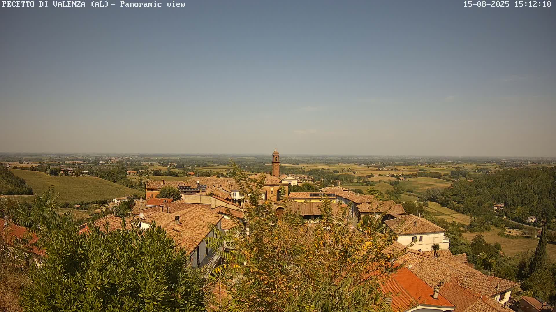 A panoramic view shows a small town with red-tiled roofs nestled in a hilly landscape under a clear, sunny sky.