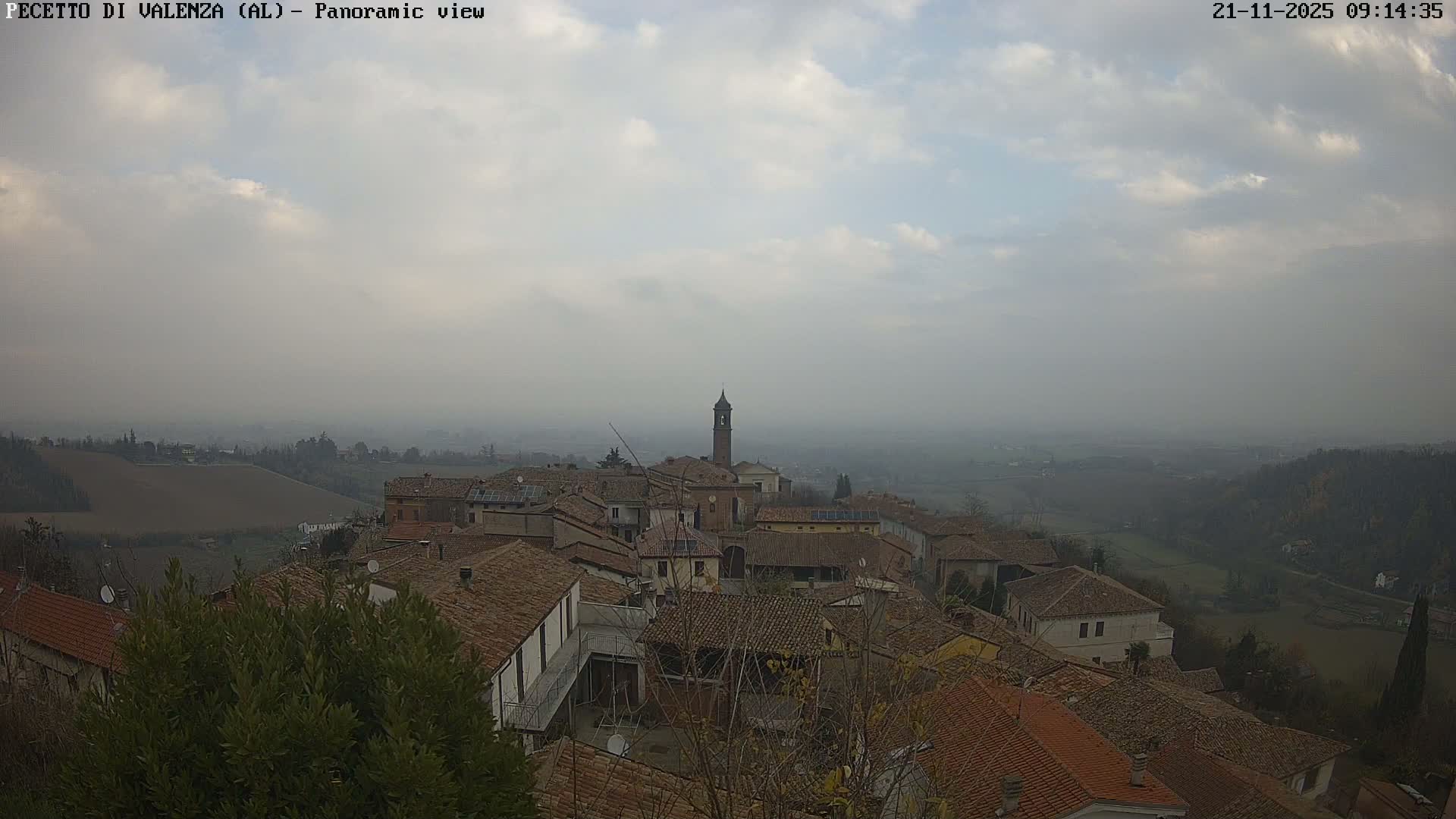 An elevated panoramic view reveals a quaint European village featuring terracotta-roofed buildings and a prominent bell tower, surrounded by rolling hills and fields under a hazy, overcast sky.